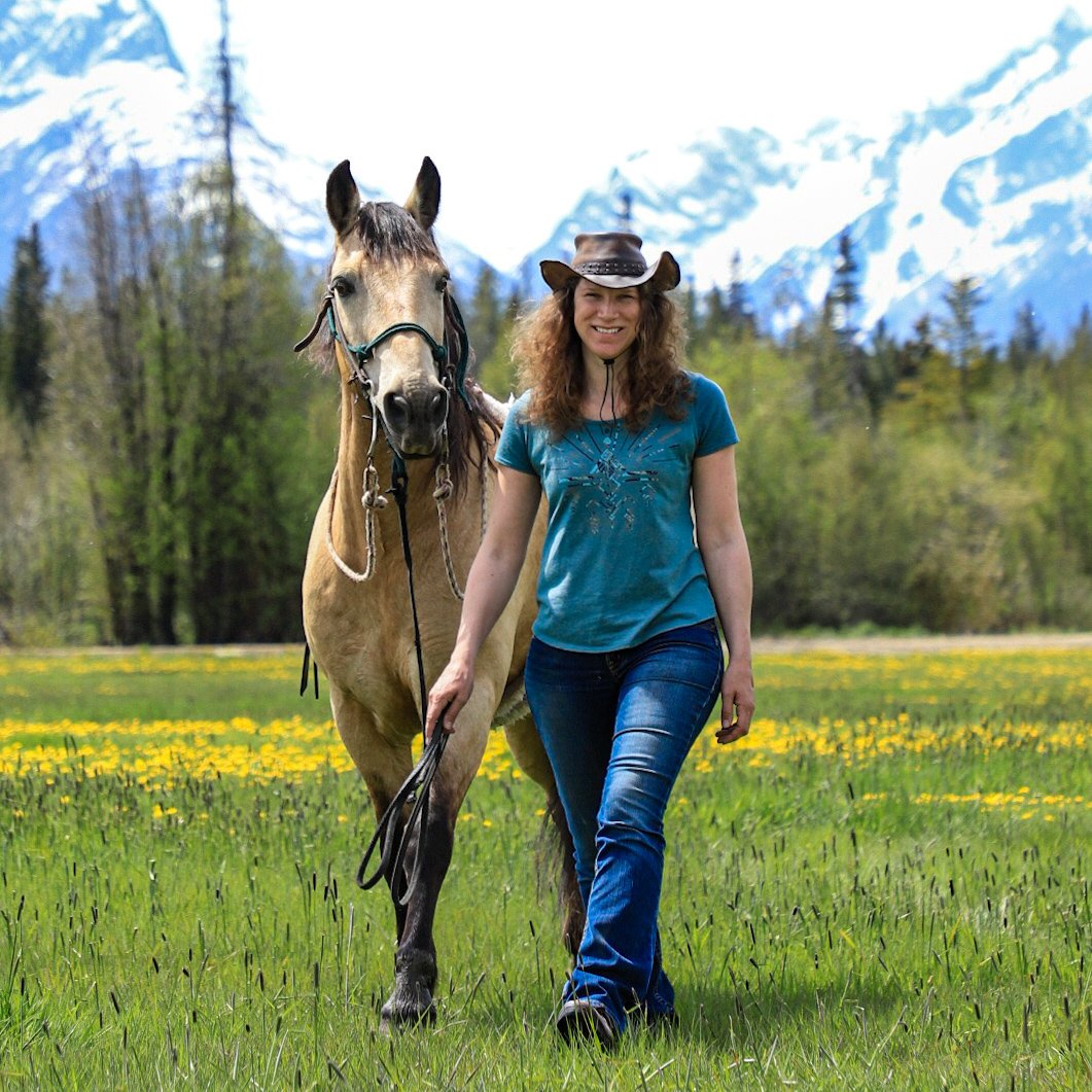 Zoe Dorhout riding a horse through a dense forest of evergreen trees with mountains in the background.