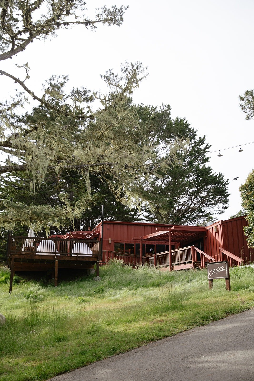 A red wooden building with an outdoor deck and stairs, surrounded by green grass and large trees draped with moss, under an overcast sky.