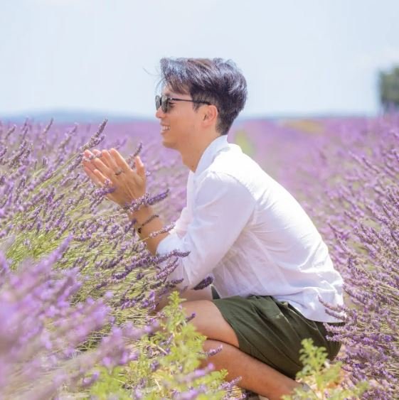 Yusuke Masuda, parfumeur, accroupi dans un champ de lavande en fleurs à Grasse, souriant et portant des lunettes de soleil, un haut blanc et un short vert