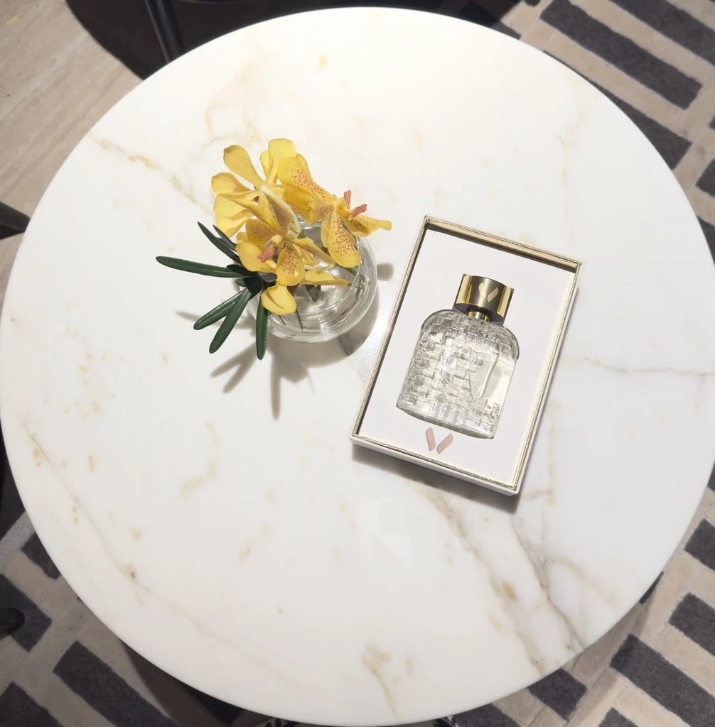 Une petite table ronde en marbre blanc avec veines dorées, située dans le hall de l'hôtel Montien à Bangkok, cette table contenant un vase avec des fleurs jaunes et un parfum dans un écrin blanc avec une bouteille en verre et un bouchon or.