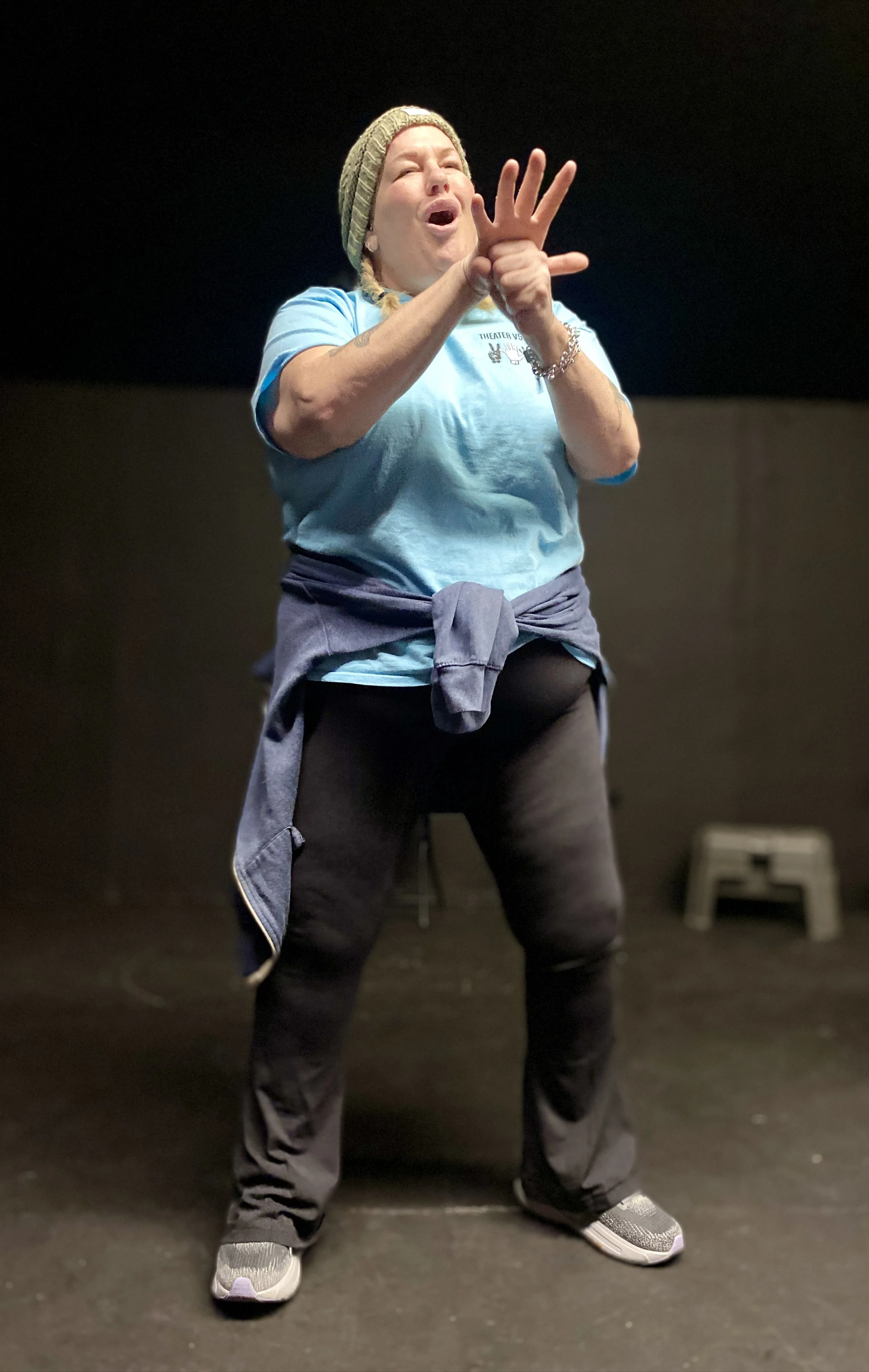A woman with blond braids and wearing a knit beanie, blue t-shirt and black jeans performs in ASL as she looks upward with her eyes closed.
