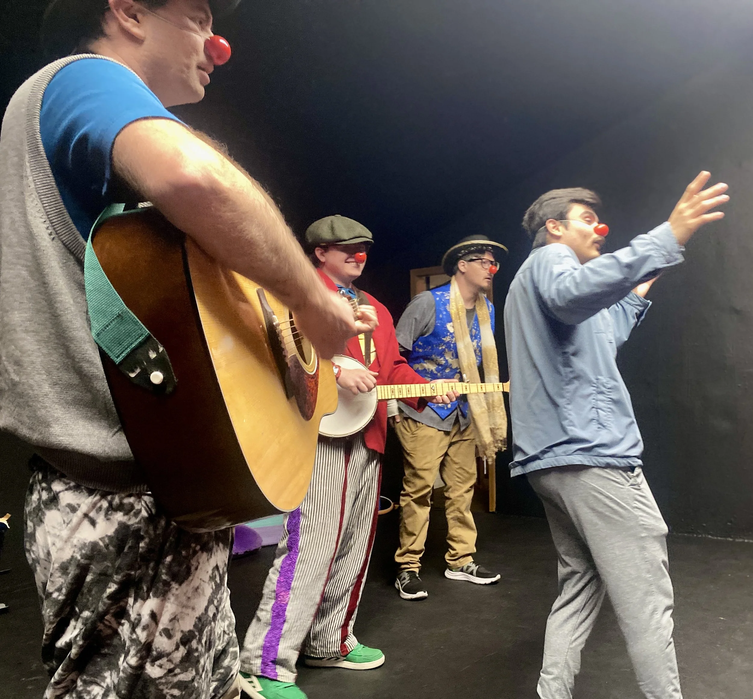 Four clowns with red noses perform music, including one guitarist wearing a blue t-shirt, one banjo player with a red jacket and bright green shoes, a singer with a blue vest and a singer wearing a blue jacket with his arms raised.