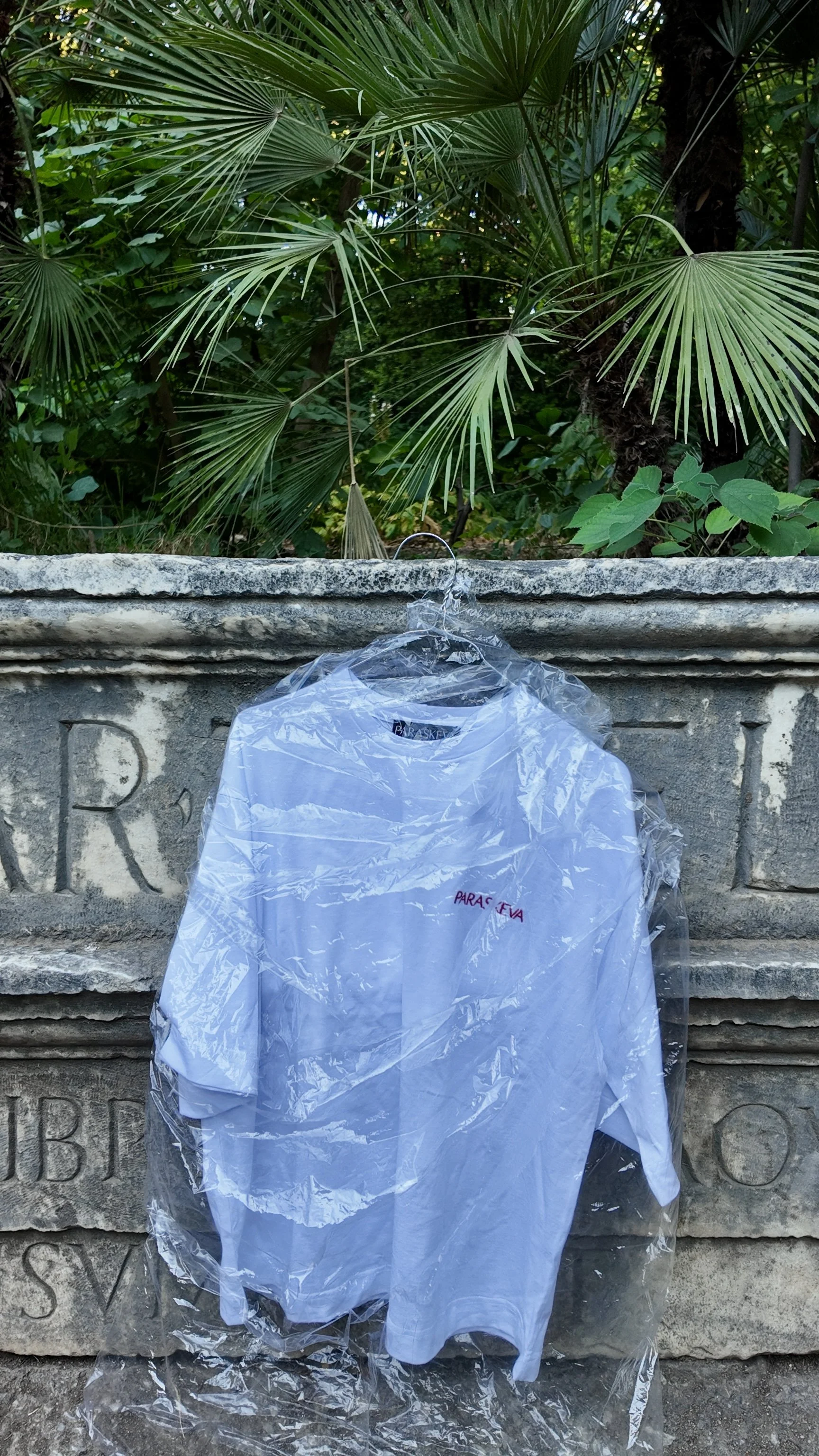 A white t-shirt in a plastic bag hanging on a metal hook in front of an old stone wall with green foliage and palm leaves in the background.
