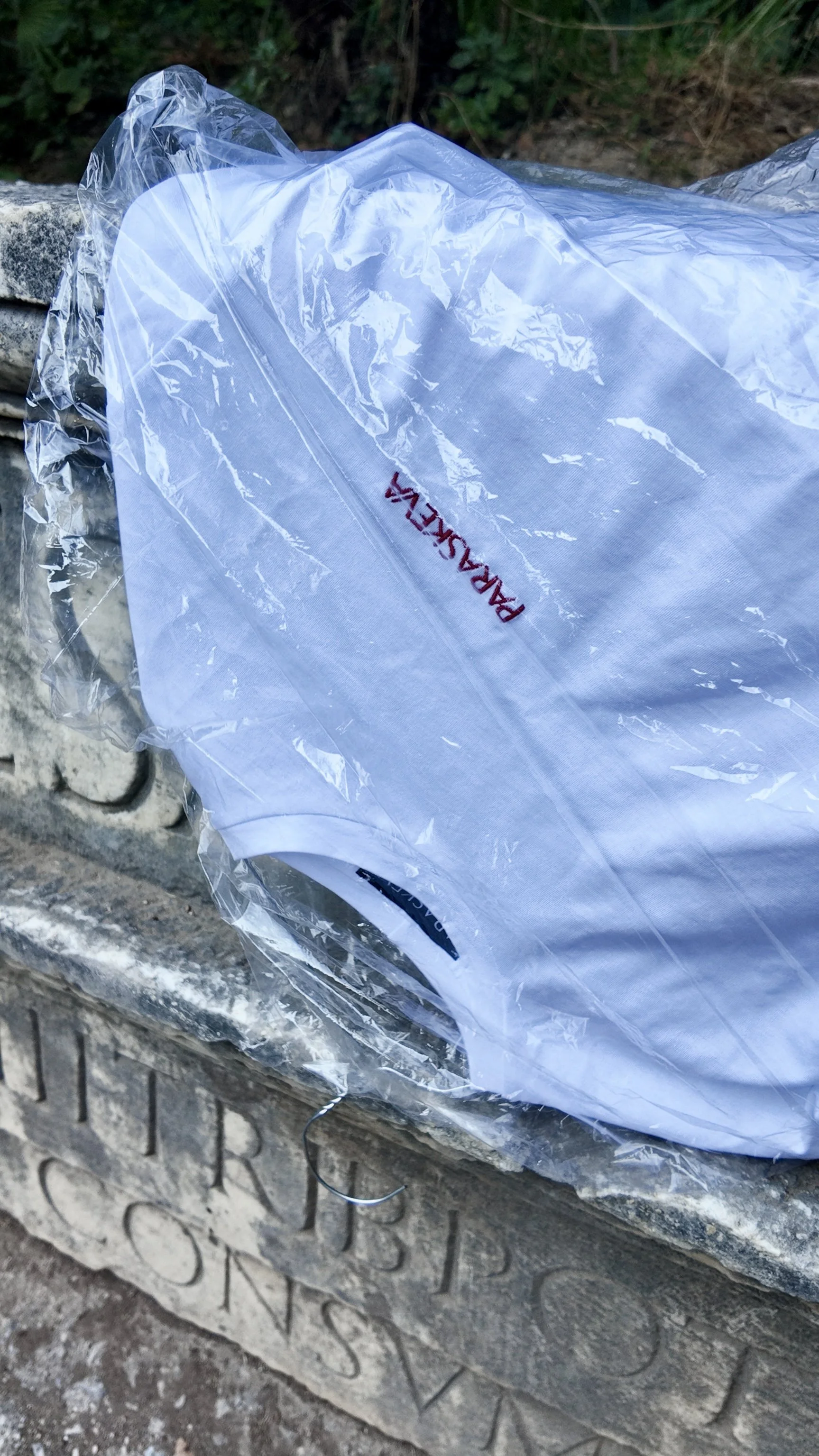 A white t-shirt with red embroidery resting inside a clear plastic bag, placed on a stone bench outdoors.