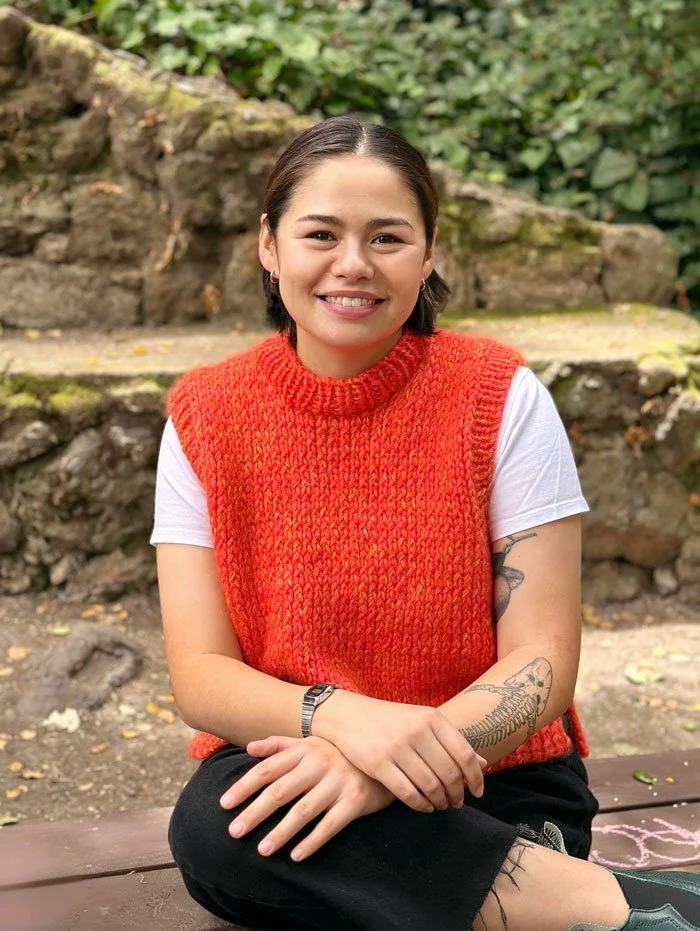Young woman sitting outdoors on a wooden bench, smiling, wearing a red knitted vest over a white T-shirt, with tattoos on her right forearm, in front of a stone wall and greenery.