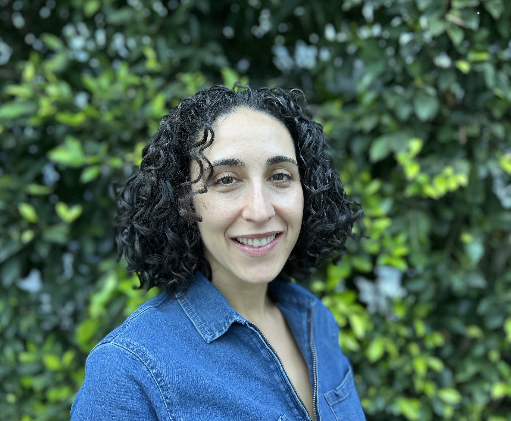 A woman with curly black hair smiling outdoors in front of green shrubbery, wearing a blue denim shirt.