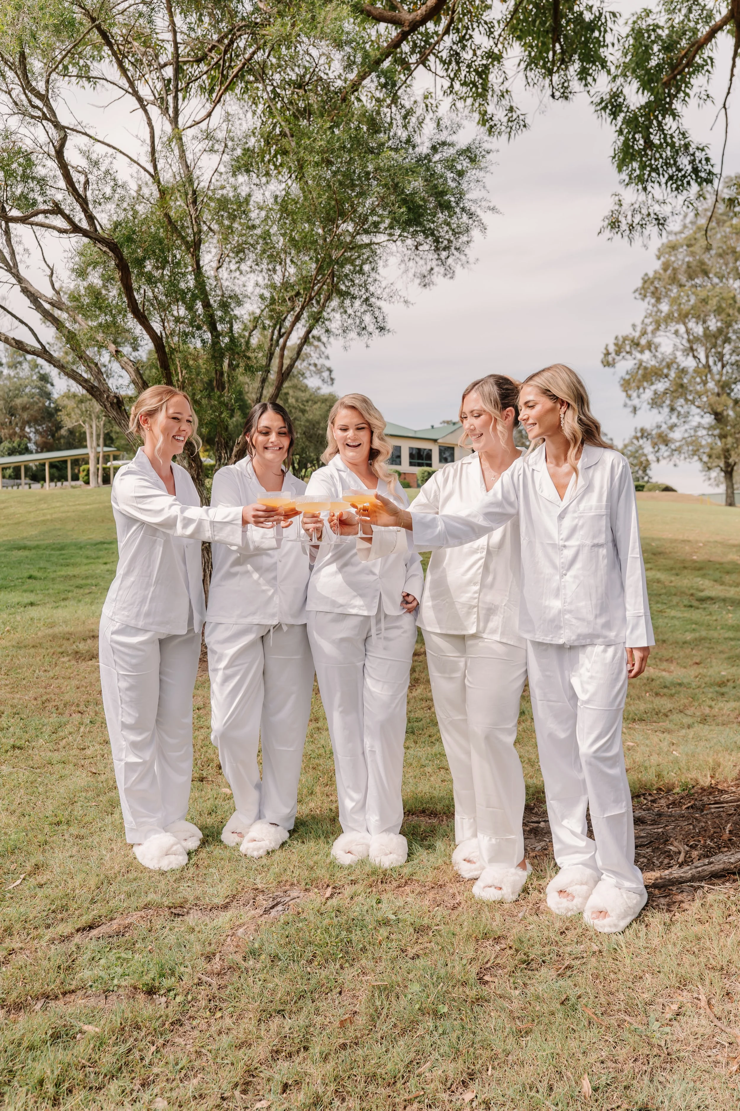 Five women dressed in white pajamas and bunny slippers standing outdoors, toasting with drinks and smiling.