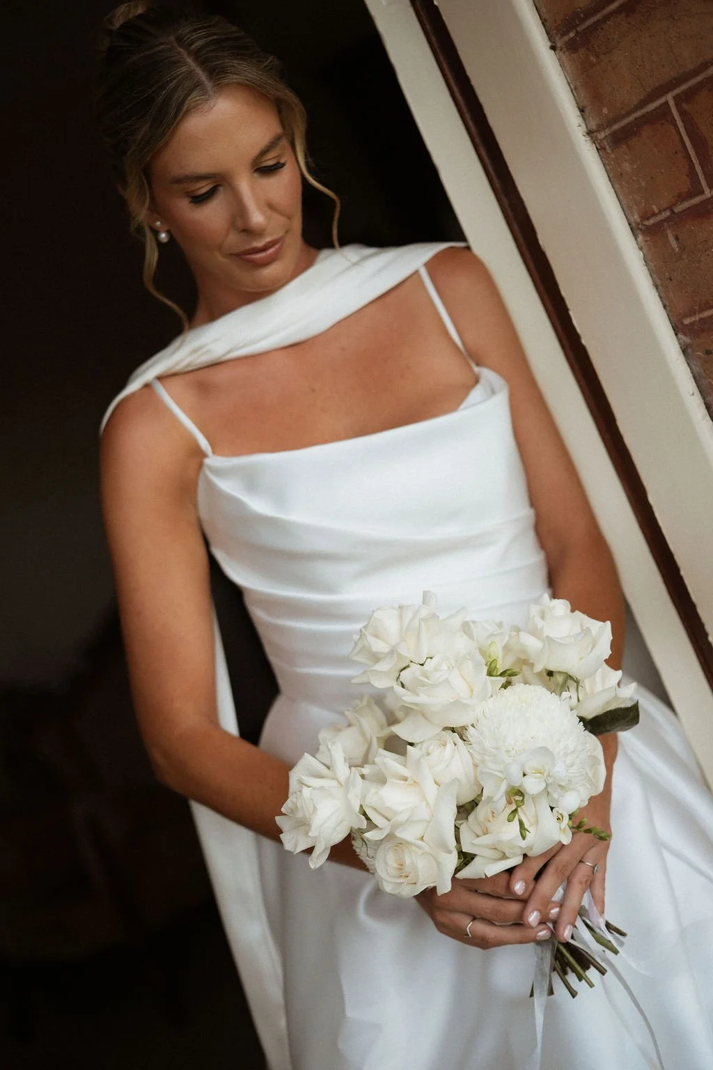 A bride in a white wedding dress holding a bouquet of white flowers, standing indoors near a brick wall.