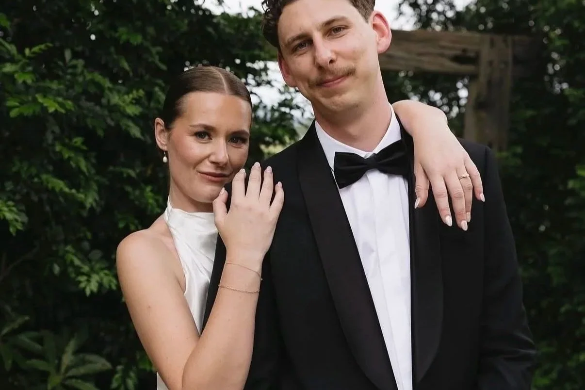 A woman and man dressed in formal wedding attire posing outdoors with greenery in the background. They are smiling with their arms around each other.