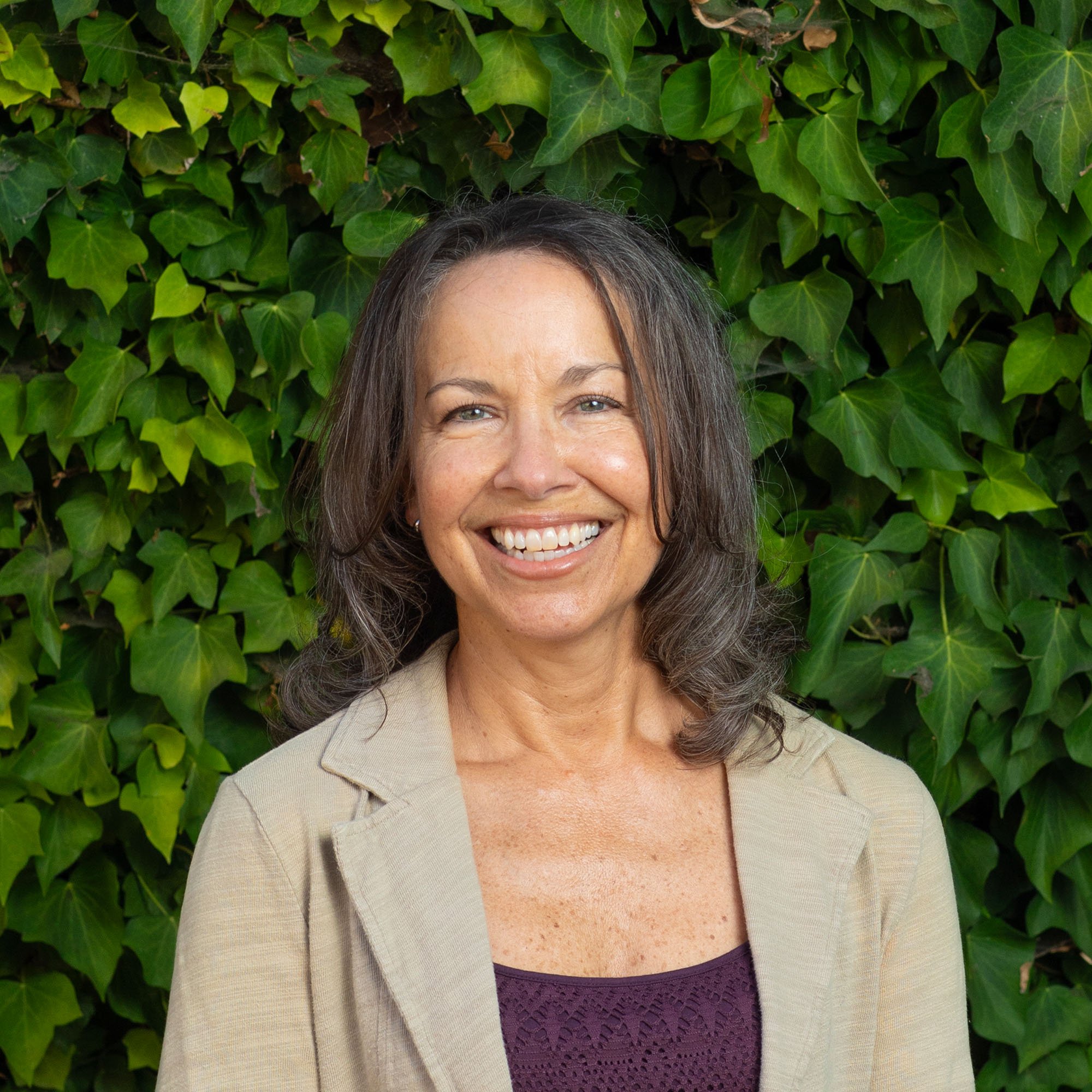 A woman with shoulder-length brown hair smiling in front of green ivy leaves.