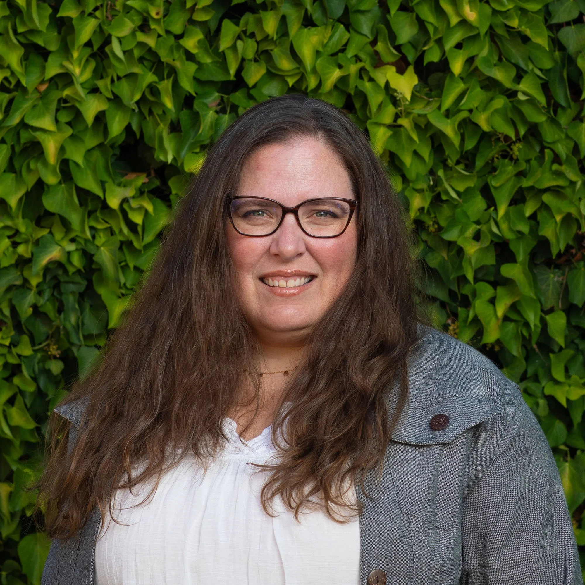 A woman with glasses and long curly brown hair smiling in front of a leafy green background.
