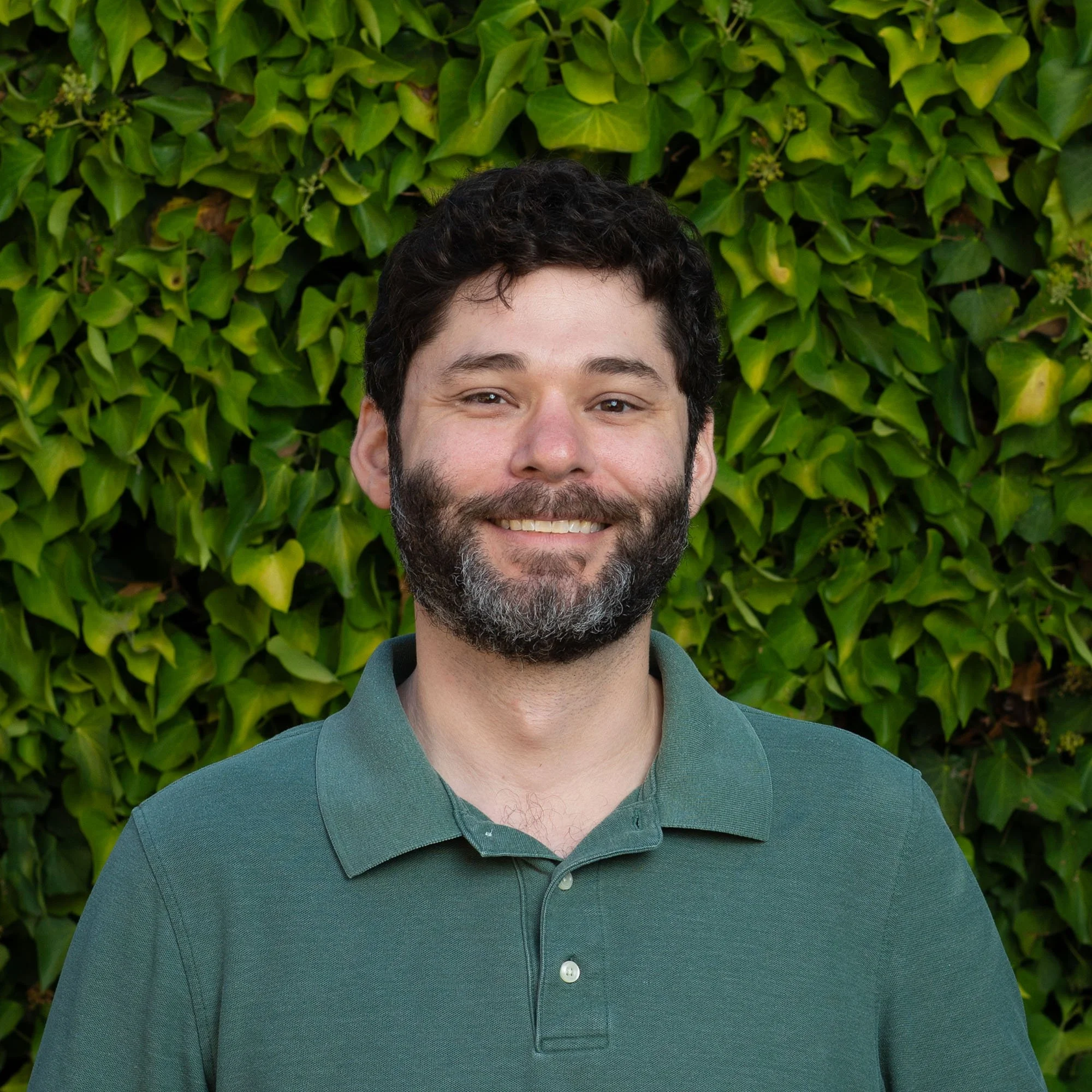 Smiling man with dark hair and beard standing in front of green leafy background.
