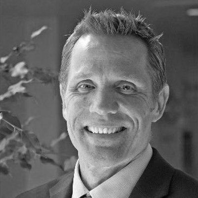 Black and white photo of a smiling man with short hair, wearing a suit and tie, standing indoors with plants in the background.