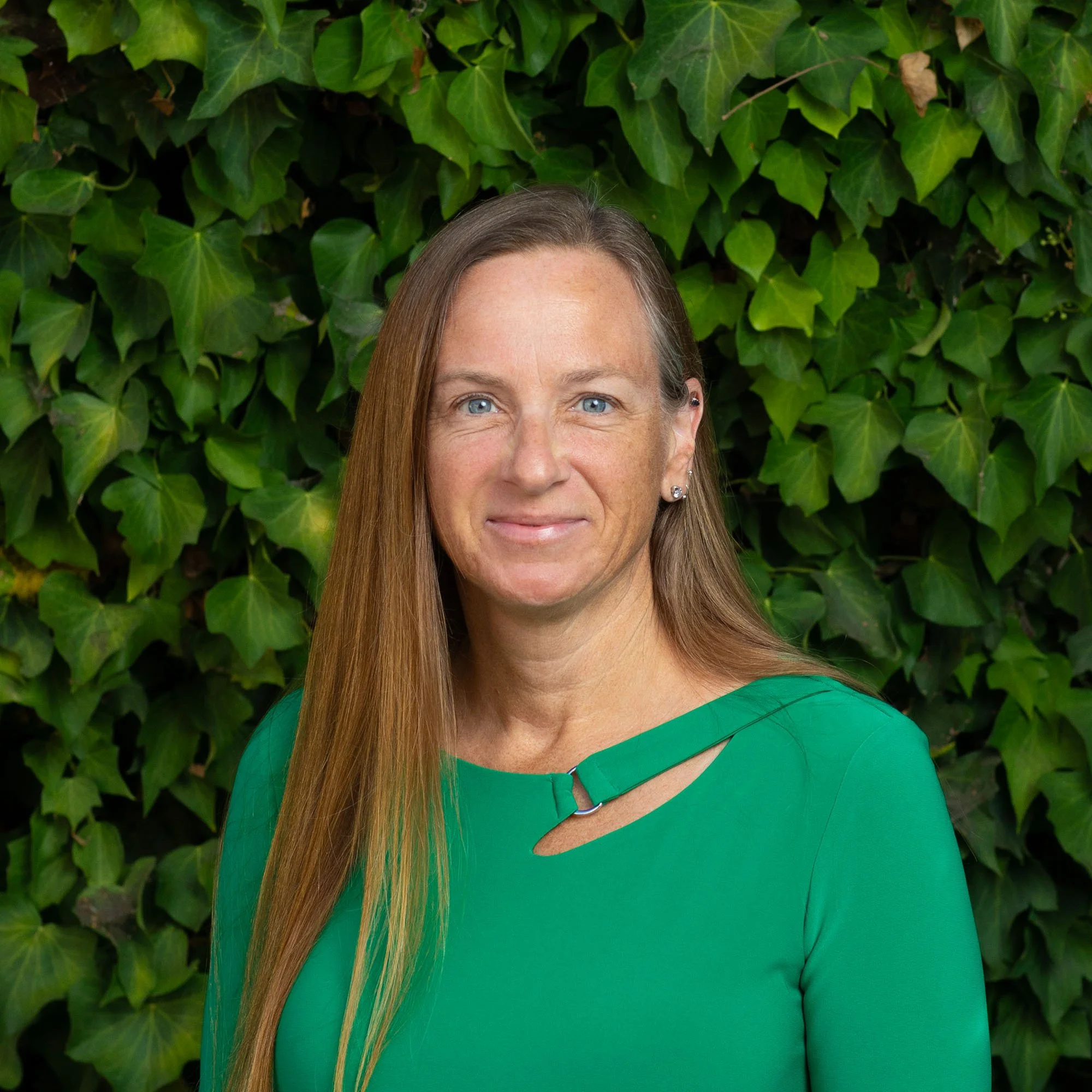 A woman with long, straight, light brown hair and blue eyes, wearing a green top with a cutout and silver jewelry, standing in front of a hedge of green leaves.