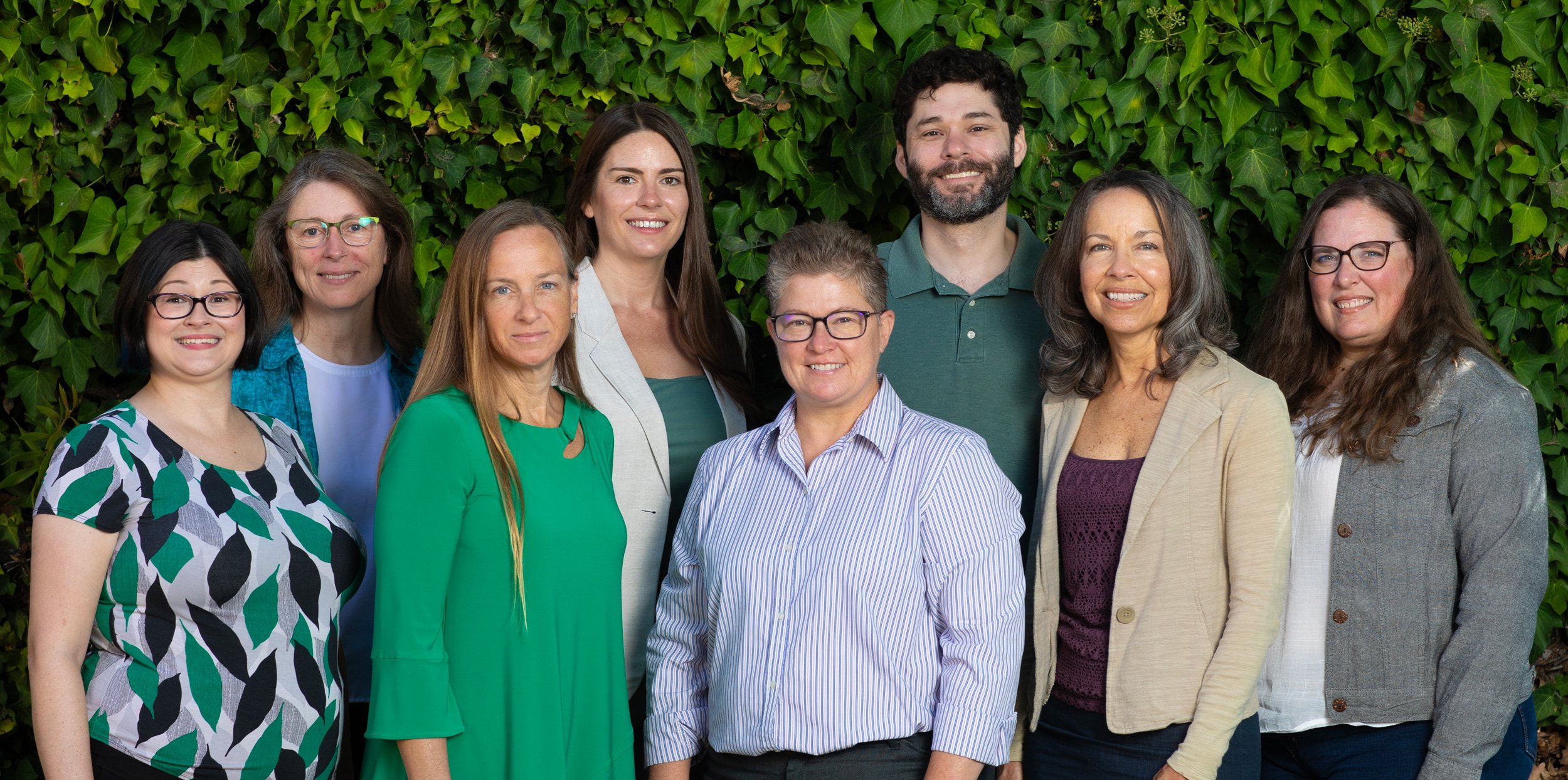 Group of nine diverse adults standing outdoors in front of green leafy hedge, smiling at camera.