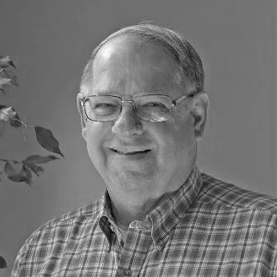 A smiling older man wearing glasses and a checkered shirt, standing indoors next to a plant.