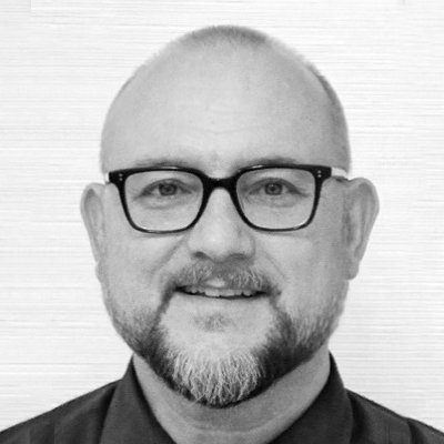 A black and white portrait photograph of a middle-aged man with glasses, short hair, and a beard, smiling against a plain background.