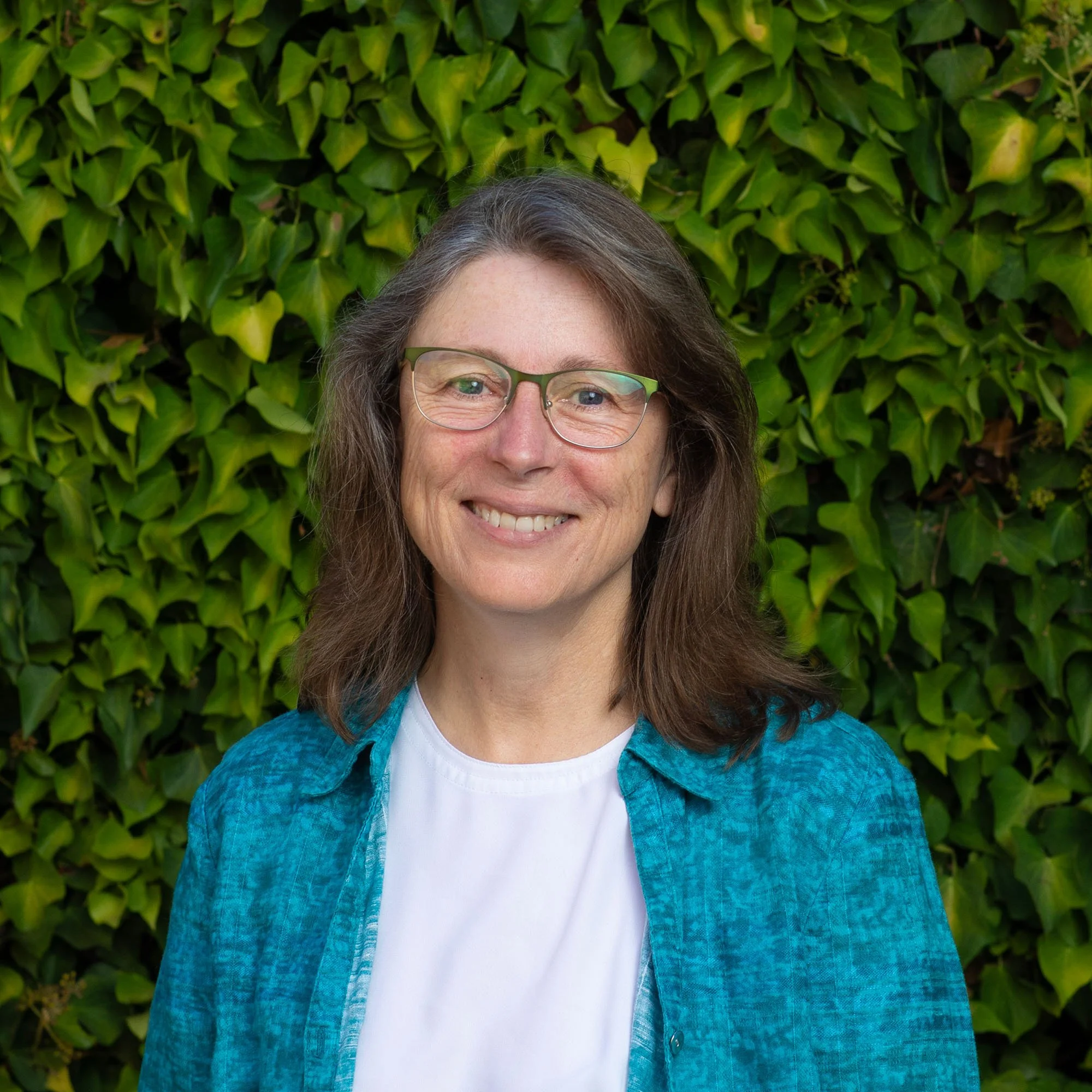 A woman with shoulder-length brown hair and glasses, smiling, wearing a white shirt and a blue plaid shirt, standing in front of green leafy bushes.