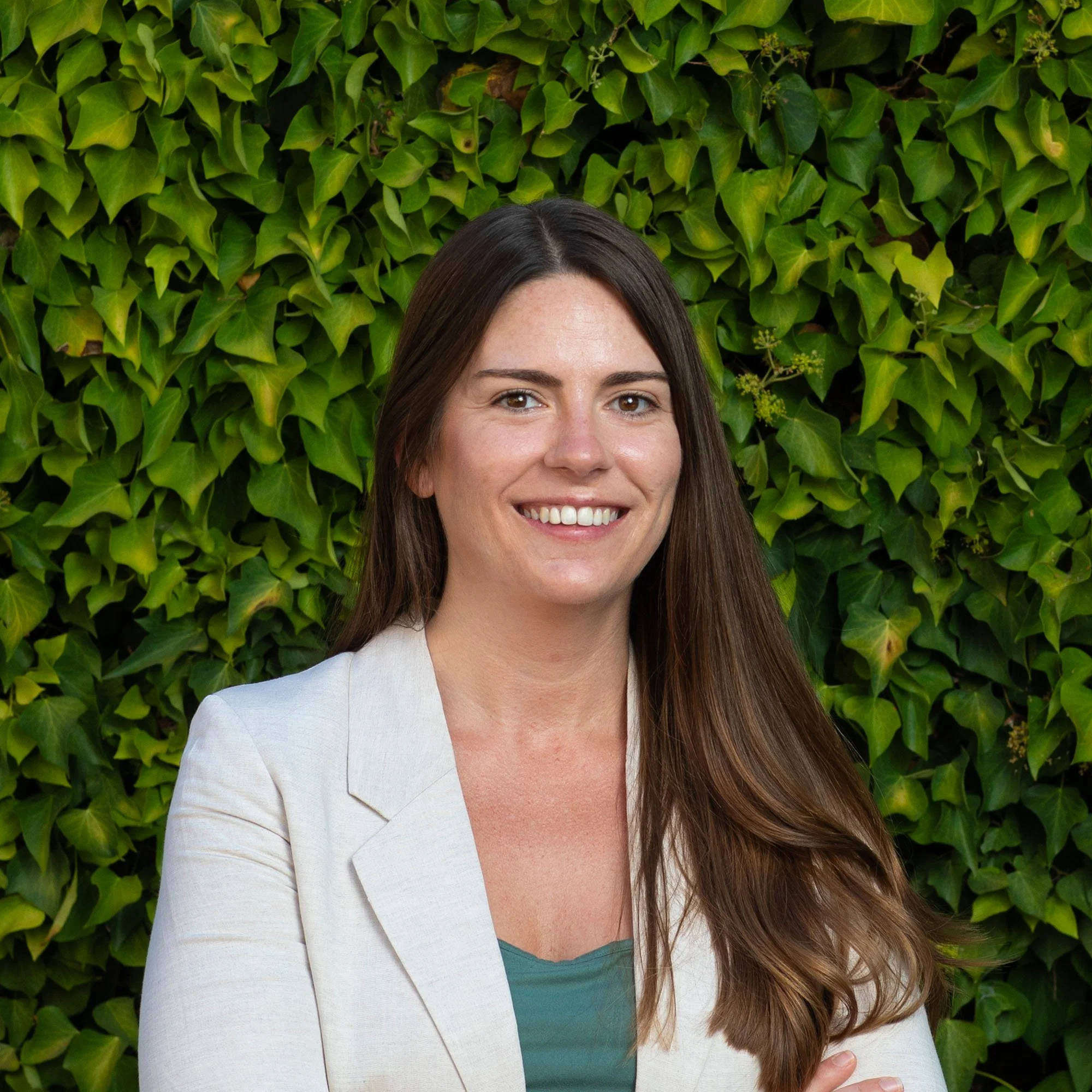 A woman with long brown hair and a white blazer standing in front of a green hedge, smiling at the camera.