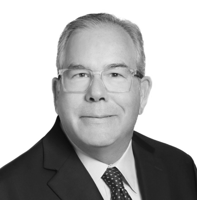 Black and white professional headshot of a middle-aged man wearing glasses, a dark suit, white shirt, and patterned tie, smiling slightly against a plain background.