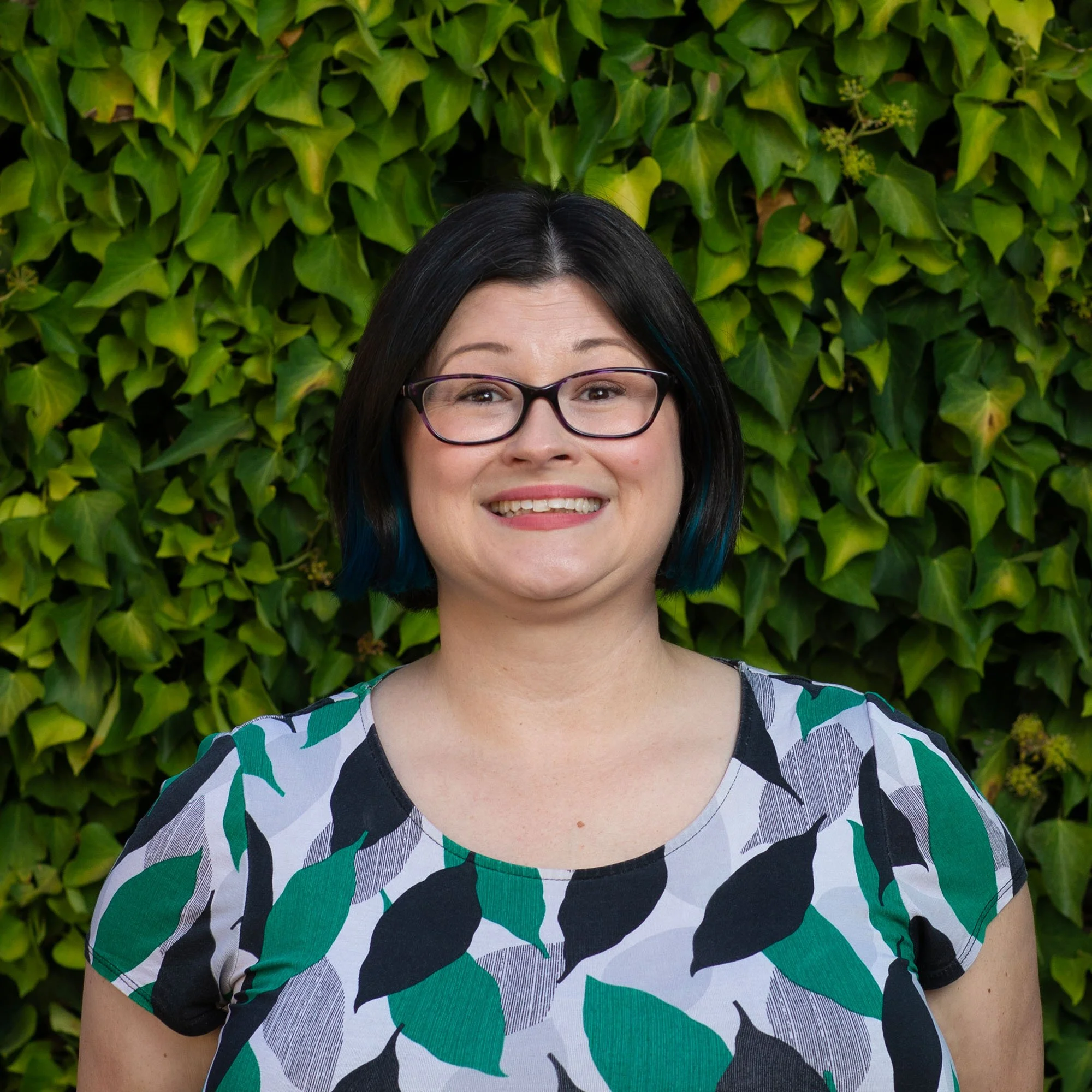 A woman with dark hair, glasses, and a patterned top standing in front of a green leafy background.