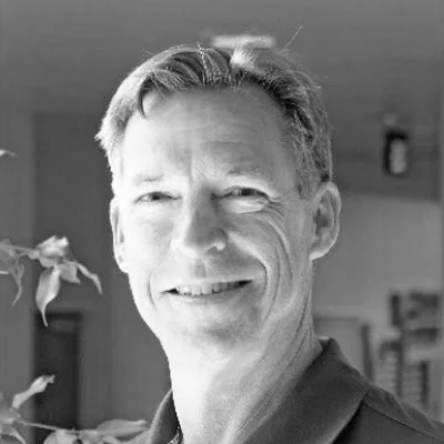 A black and white photo of a middle-aged man smiling, with short hair, indoors with a plant and some furniture visible in the background.