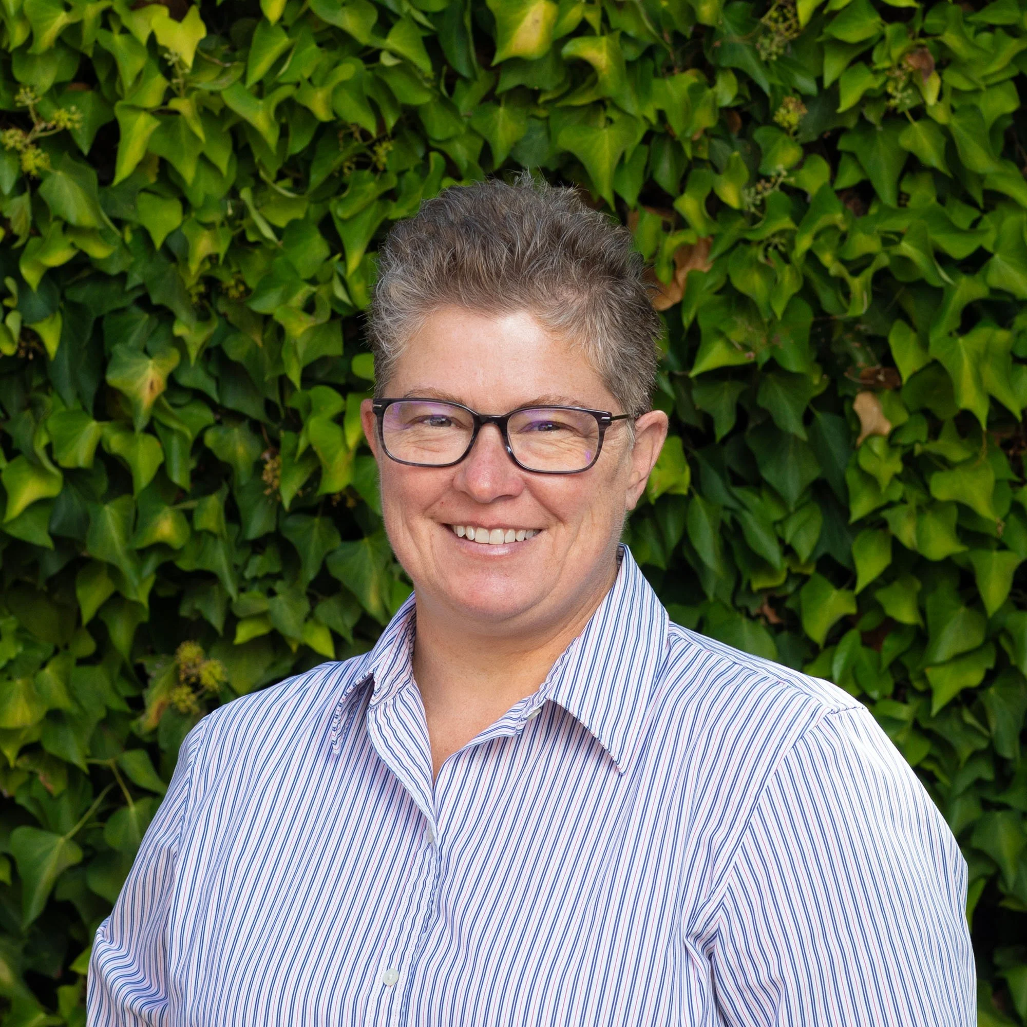 A person with short, light-colored hair and glasses smiling in front of a green leafy background.