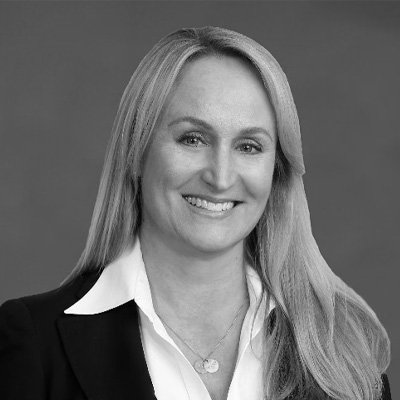 Businesswoman with long blonde hair, wearing a black blazer and white shirt, smiling, in a professional headshot.