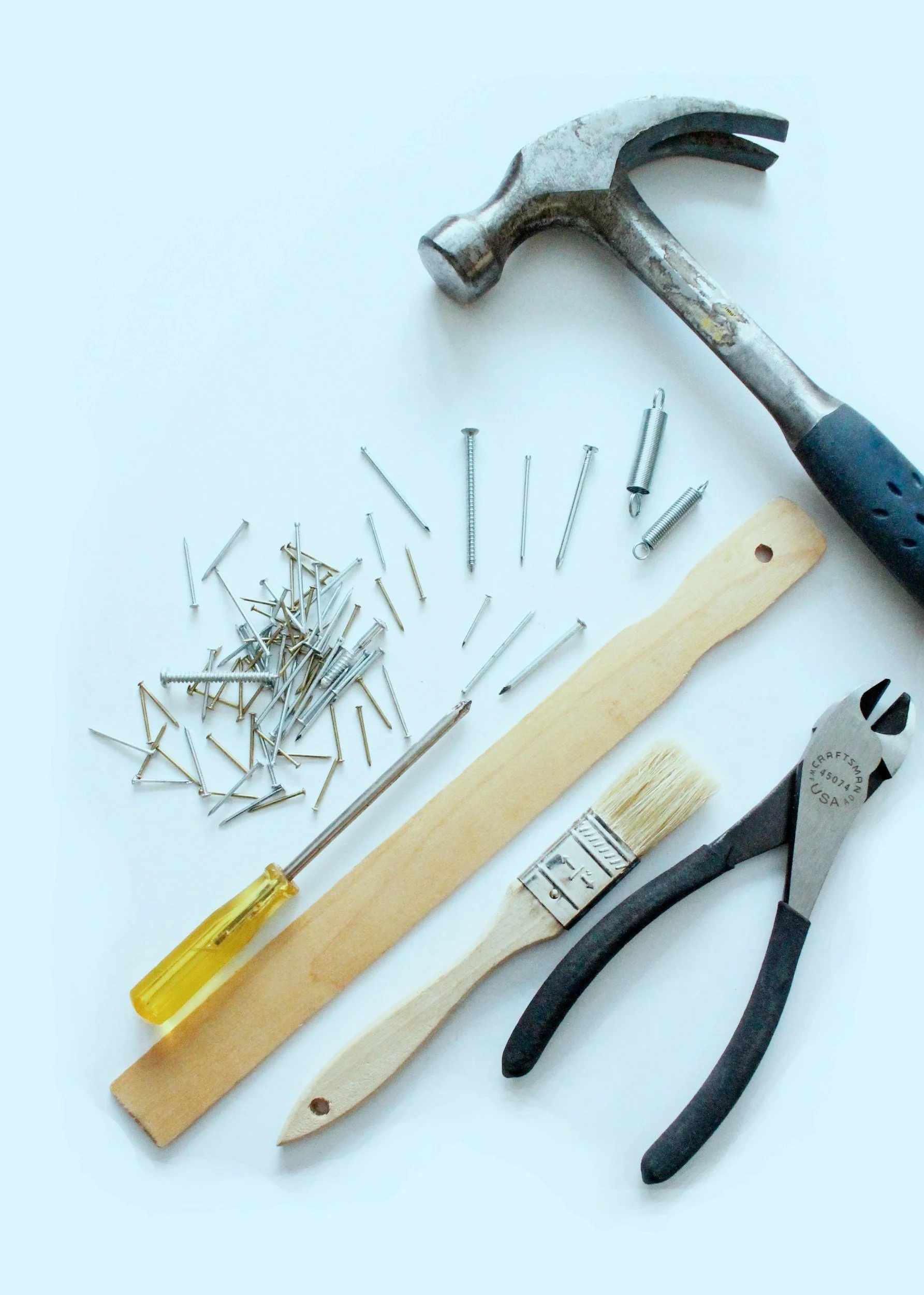 Hammer, screwdriver, nails, screws, springs, a wood stick, a paintbrush, and pliers on a white surface.