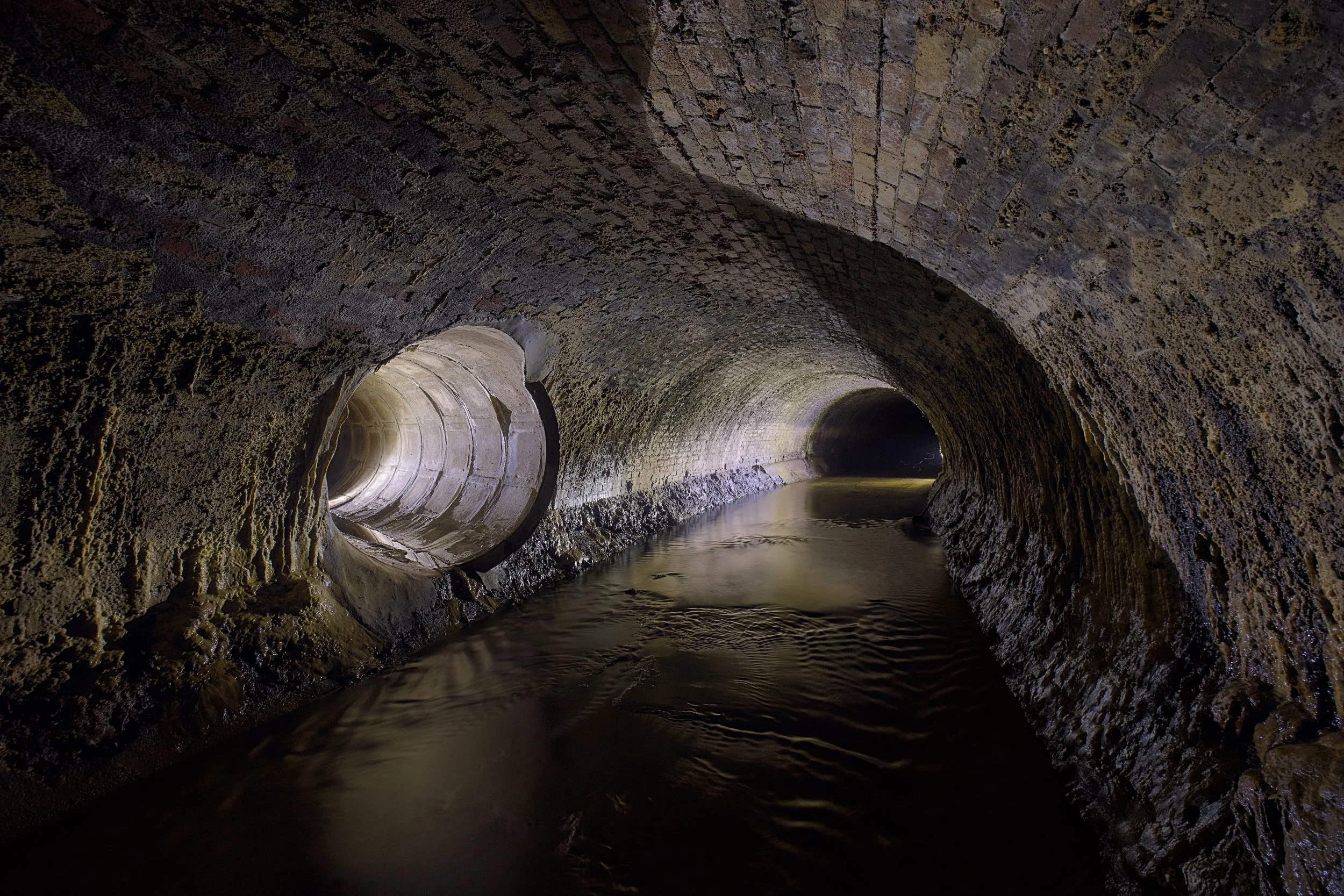 An interior shot looking down a dark, arched subterranean brick tunnel with a stream of murky water flowing through the center.
The tunnel walls are made of weathered, brown brick and stone, heavily coated with textured mineral deposits and grime. On