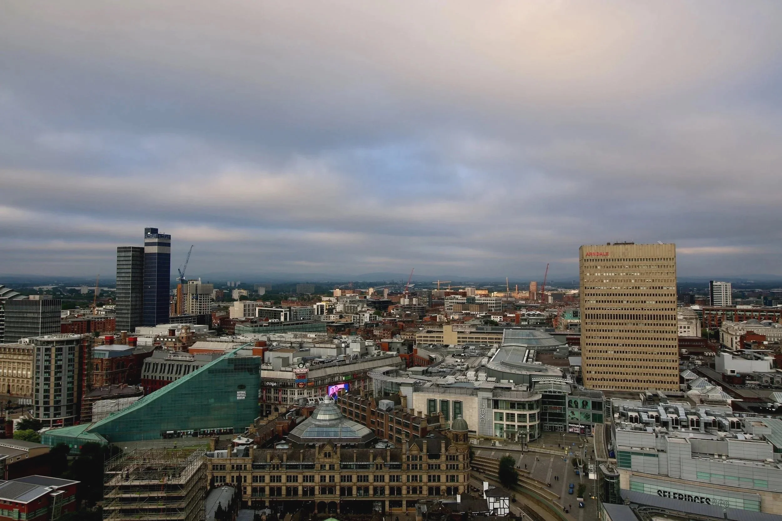 A wide view of central Manchester seen from a rooftop vantage point, with a mix of modern high-rise towers, shopping centres, and historic buildings stretching across the city. Thick grey clouds fill most of the sky, while soft light breaks through i
