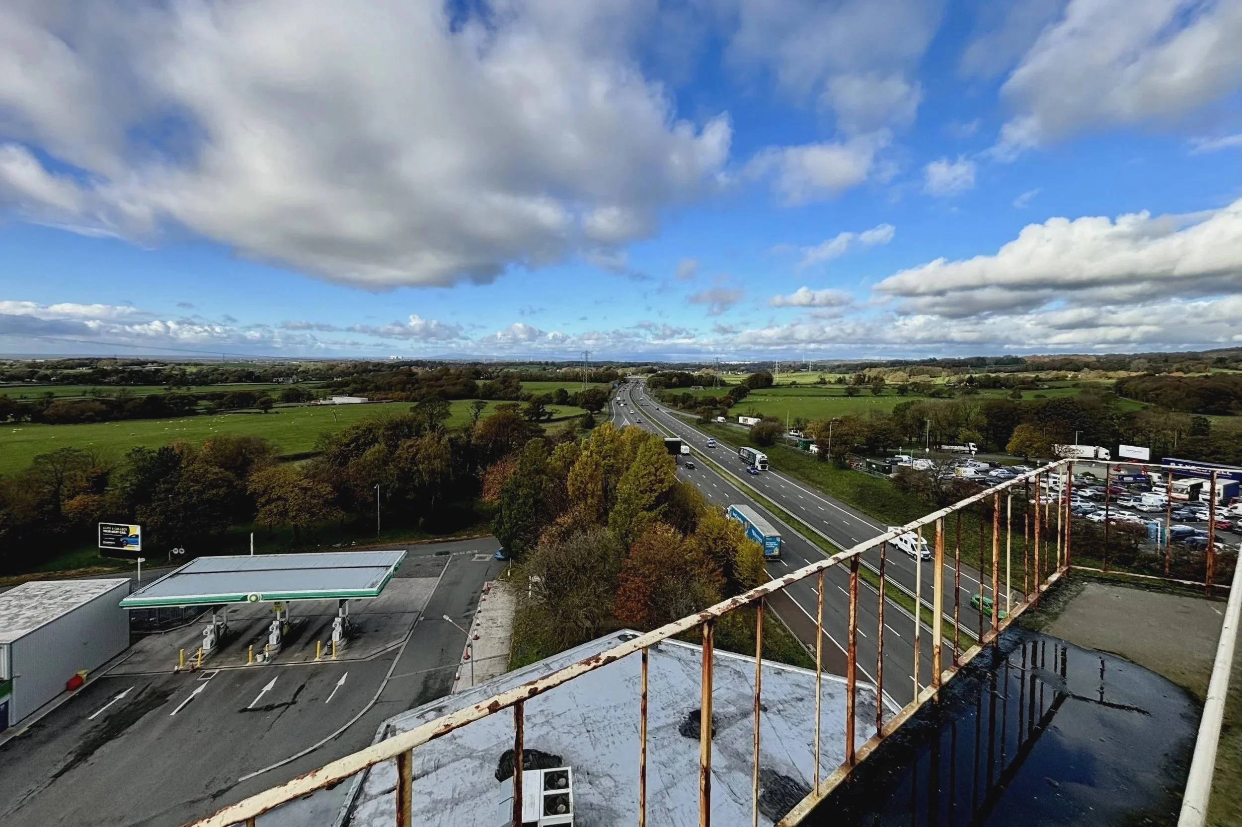 A panoramic view from the Pennine Tower overlooks the M6 motorway as it stretches through open countryside. Multiple lanes of traffic run between green fields and clusters of autumn-coloured trees, while a service station and rooftop structures appea