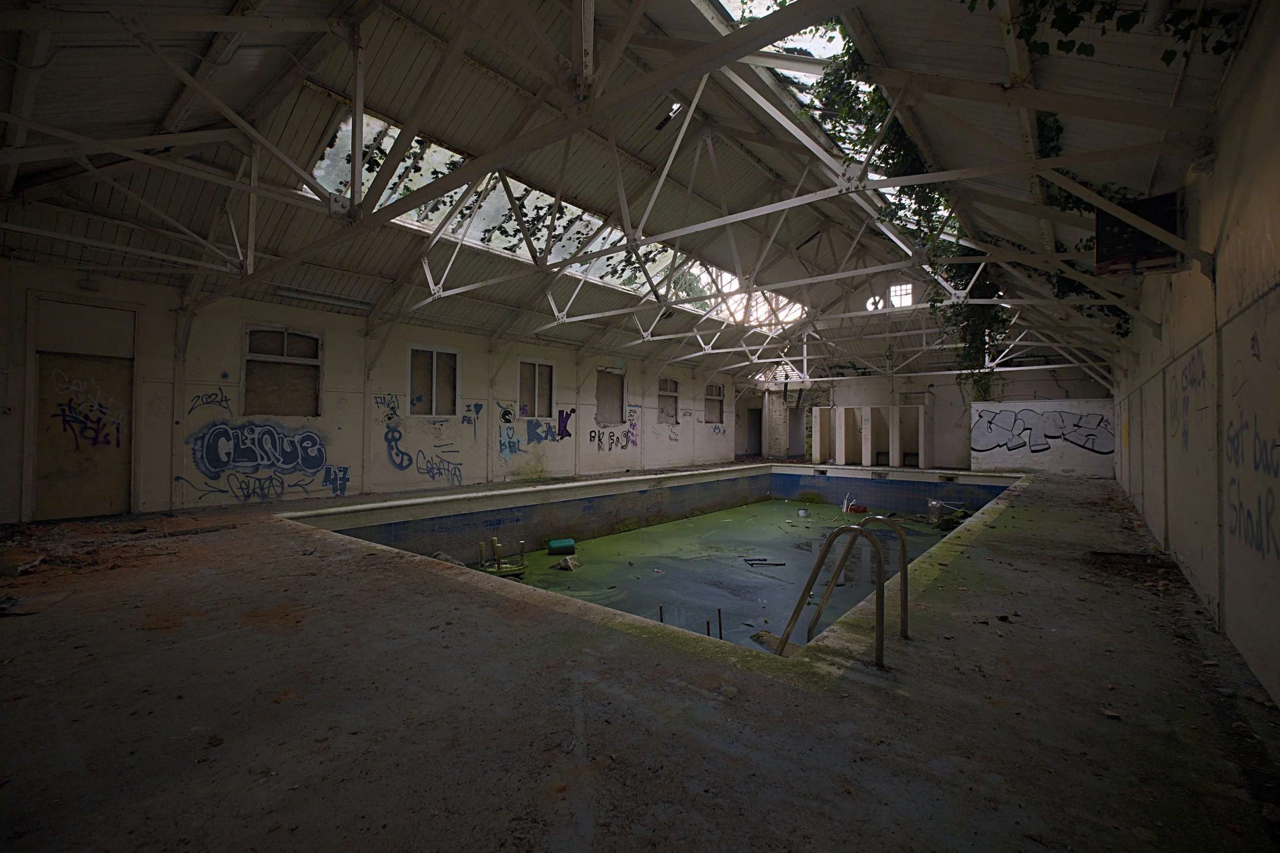 A wide-angle, interior shot of an abandoned indoor swimming pool facility.
The large, rectangular pool in the center is mostly empty of water but filled with thick, bright green algae, moss, and scattered debris. A rusted metal pool ladder stands at 
