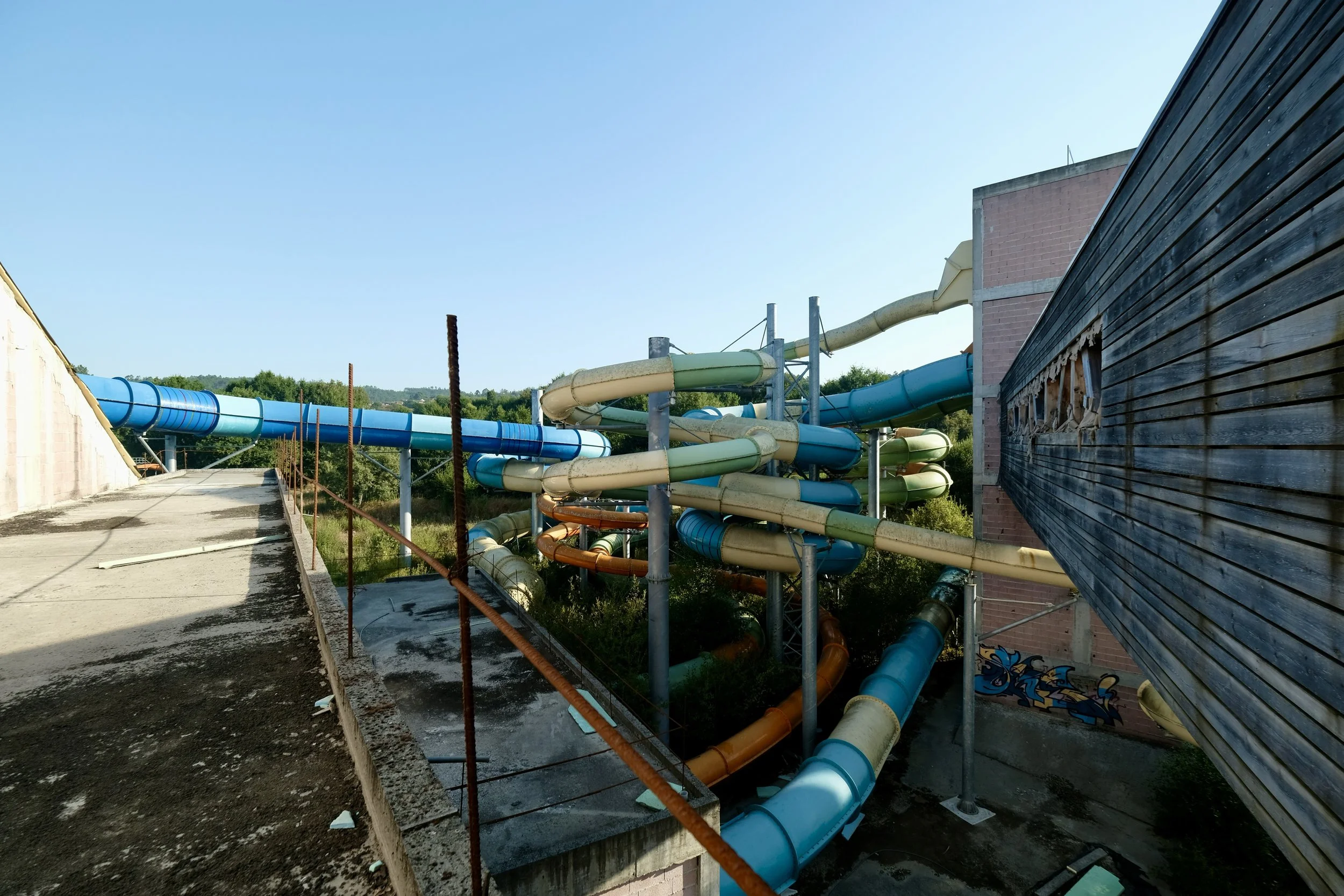 An outdoor, wide-angle shot of an abandoned or derelict water park complex, taken on a clear, sunny day. The scene is dominated by a tangled, multi-tiered network of large, colorful water slide tubes weaving between weathered concrete and wooden stru