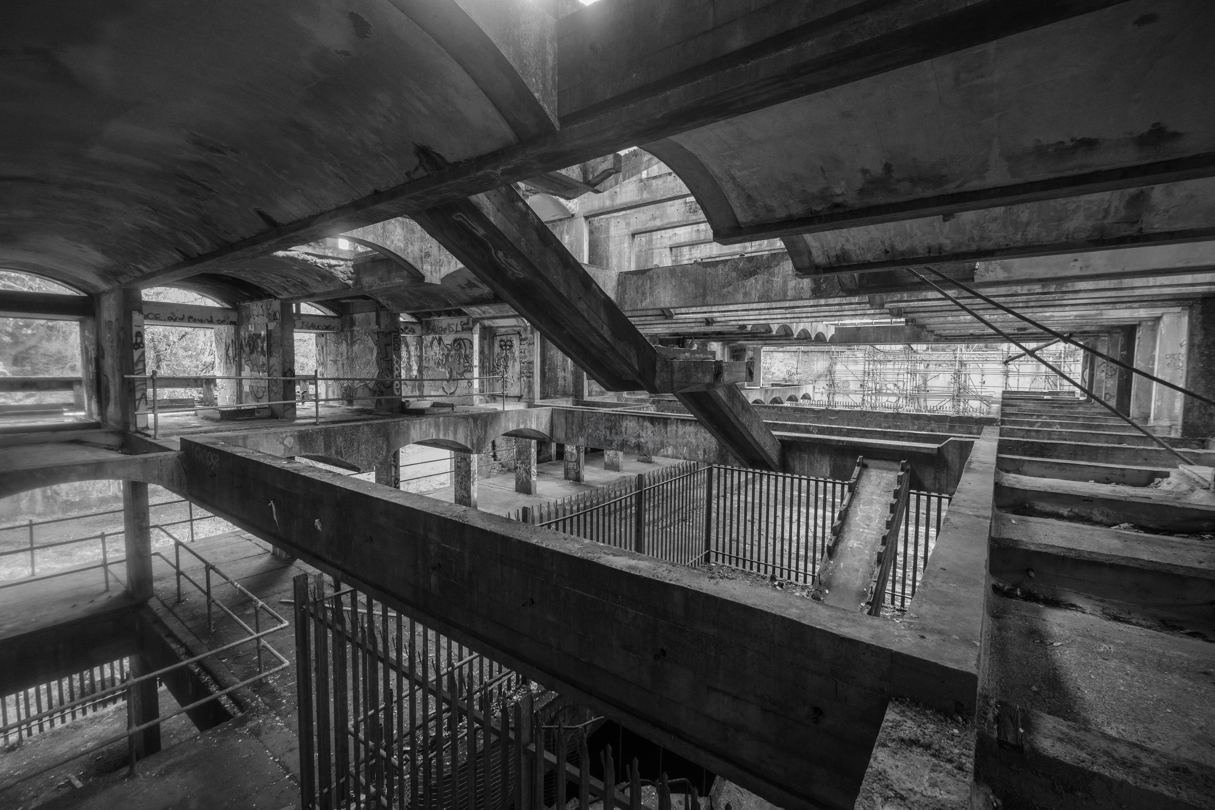 A black-and-white, wide-angle photograph capturing the interior of a massive, brutalist concrete structure left in ruins.
The image features multiple exposed levels, intersecting walkways, and stark geometric concrete beams spanning the open, caverno