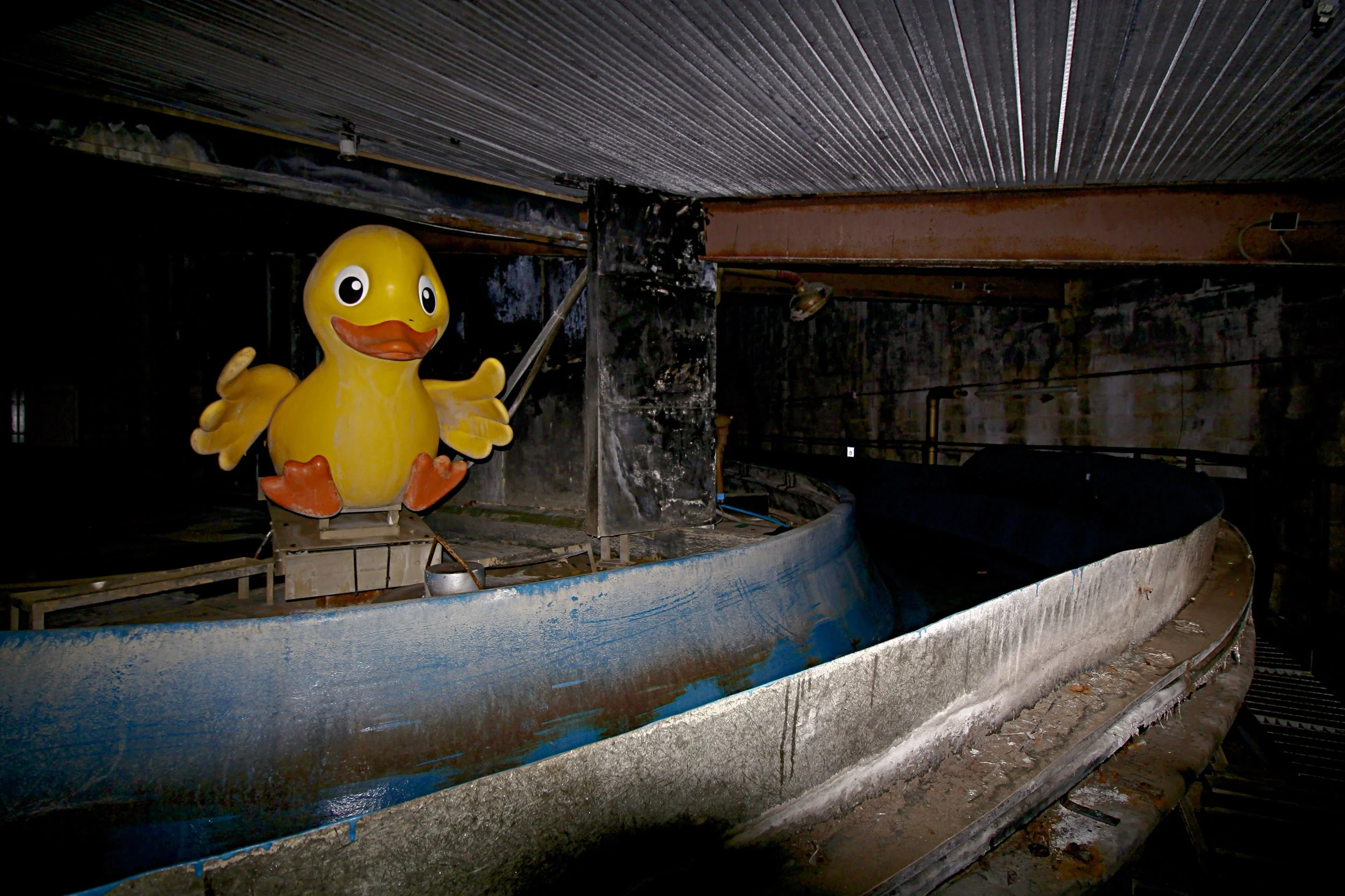 A flash-illuminated, interior shot of an abandoned, dark indoor water ride or amusement attraction.
In the foreground, a curved, empty concrete and blue fiberglass water flume channel winds through the space. Perched on a mechanical platform in the c