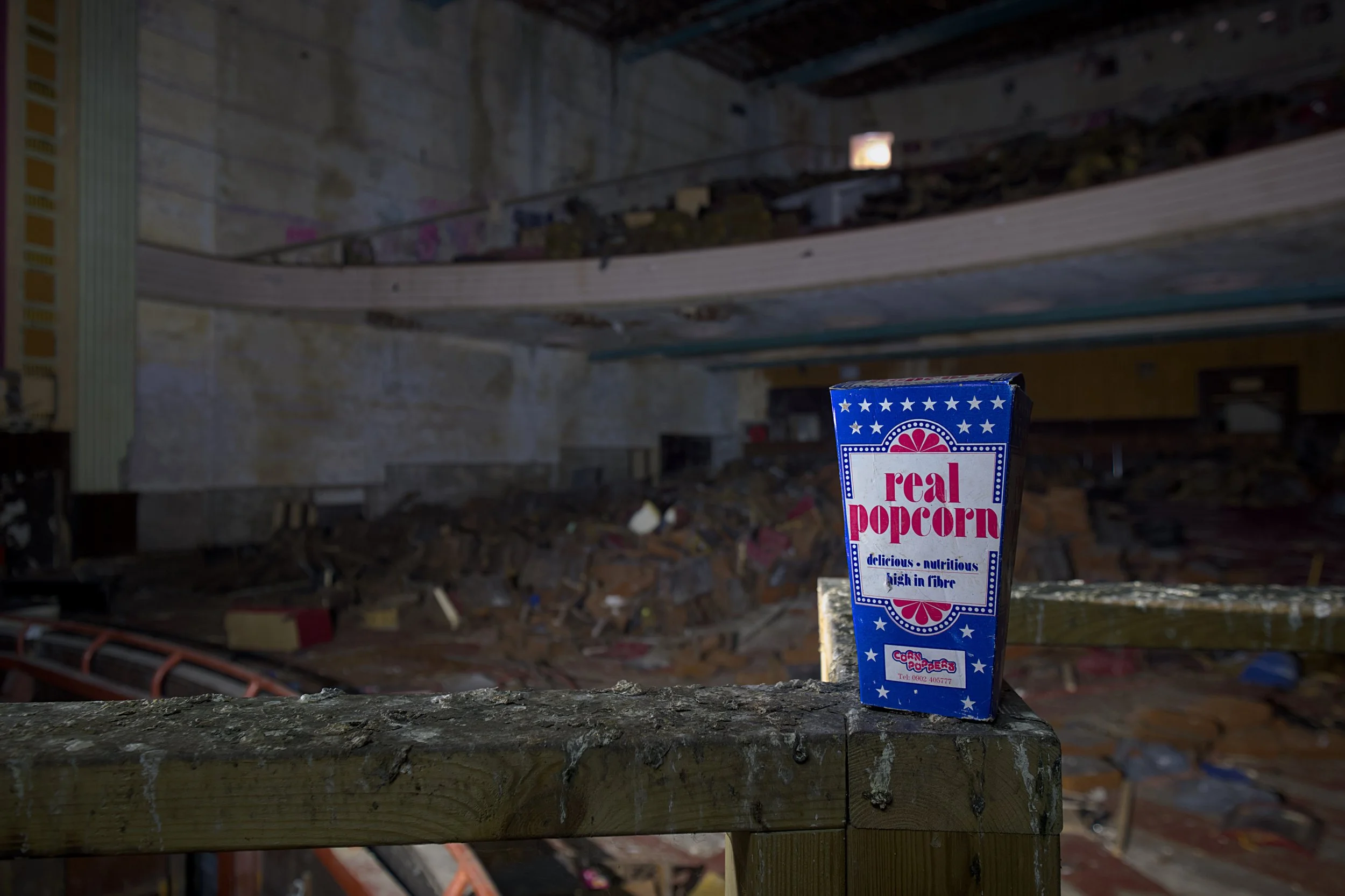 A close-up shot of a vintage, blue-and-white cardboard popcorn box sitting on a dirty, weathered wooden railing. The box features the text "real popcorn" in red letters, with smaller text below reading "delicious • nutritious • high in fibre".
The ba