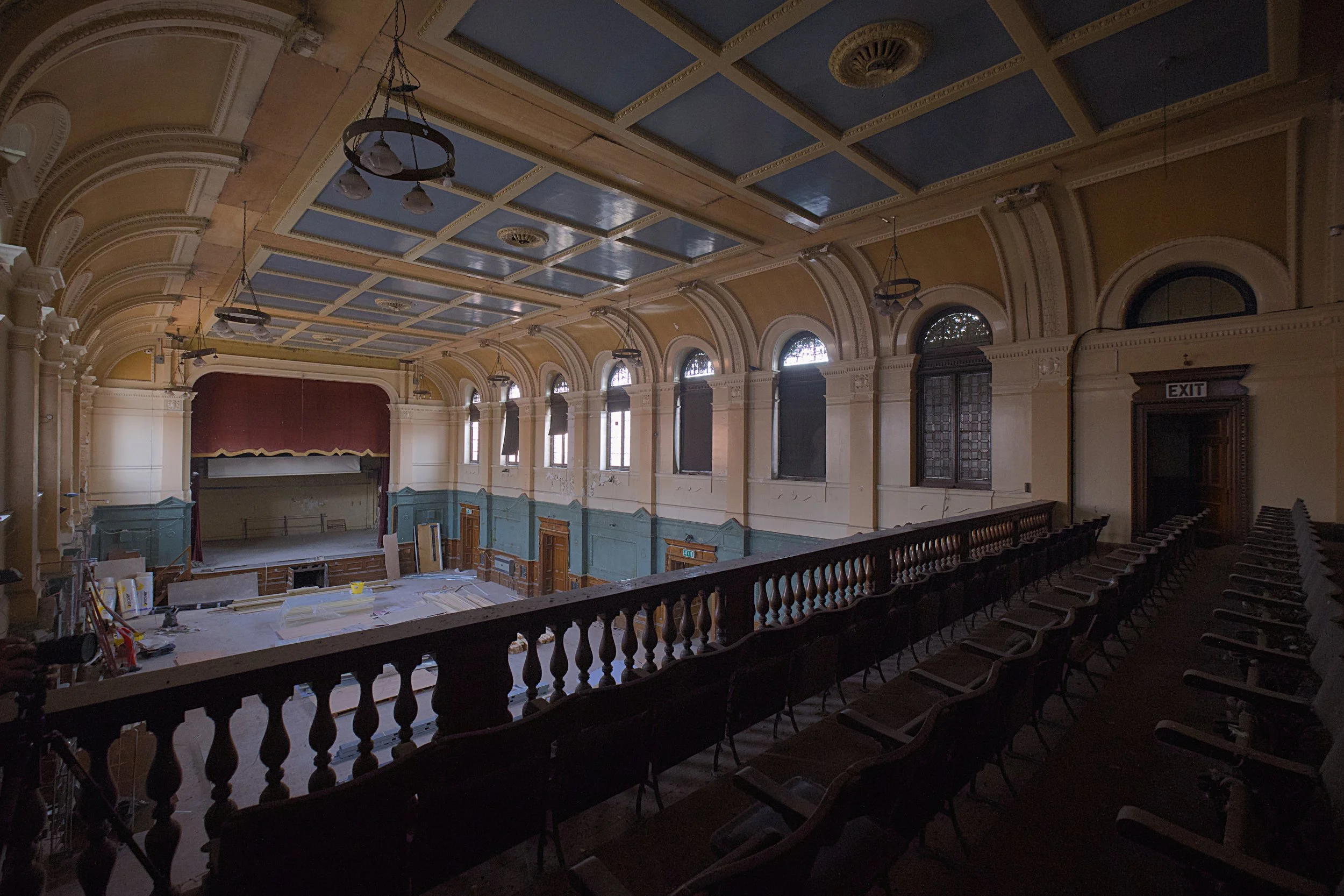 Interior view of a large historic auditorium seen from an upper balcony. Rows of seats line the balcony in the foreground, overlooking a spacious hall with a stage framed by red curtains at the far end. The room features high arched windows, decorati