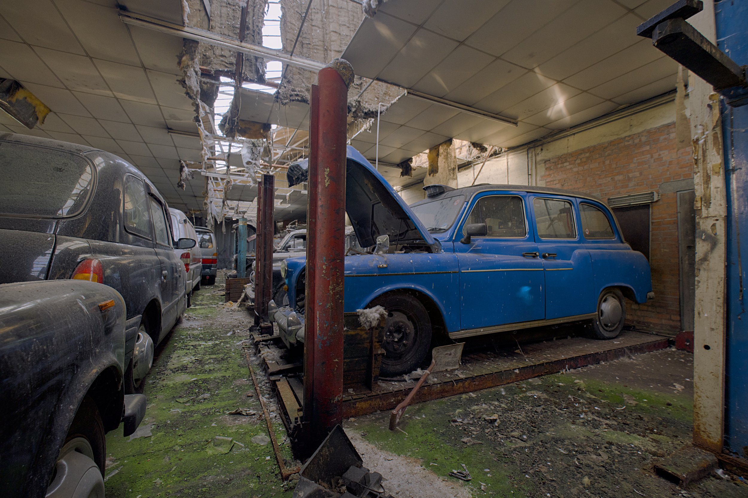 An abandoned vehicle repair garage with several dusty vintage cars parked inside. A bright blue classic car sits on a rusted lift with its bonnet open, while the damaged ceiling has large holes exposing roof beams. Debris, moss-covered concrete, and 