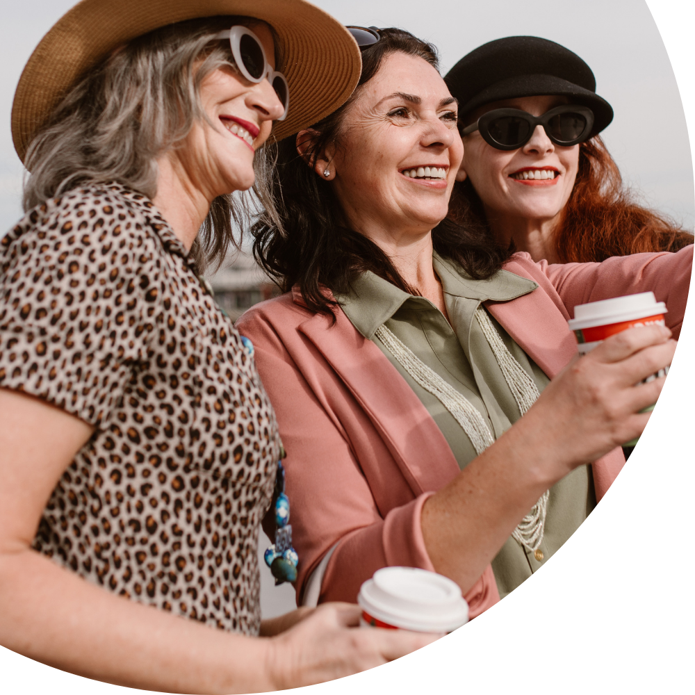 Three women outdoors, smiling and taking a selfie together, each holding a coffee cup, wearing fashionable sun hats and sunglasses, during daytime