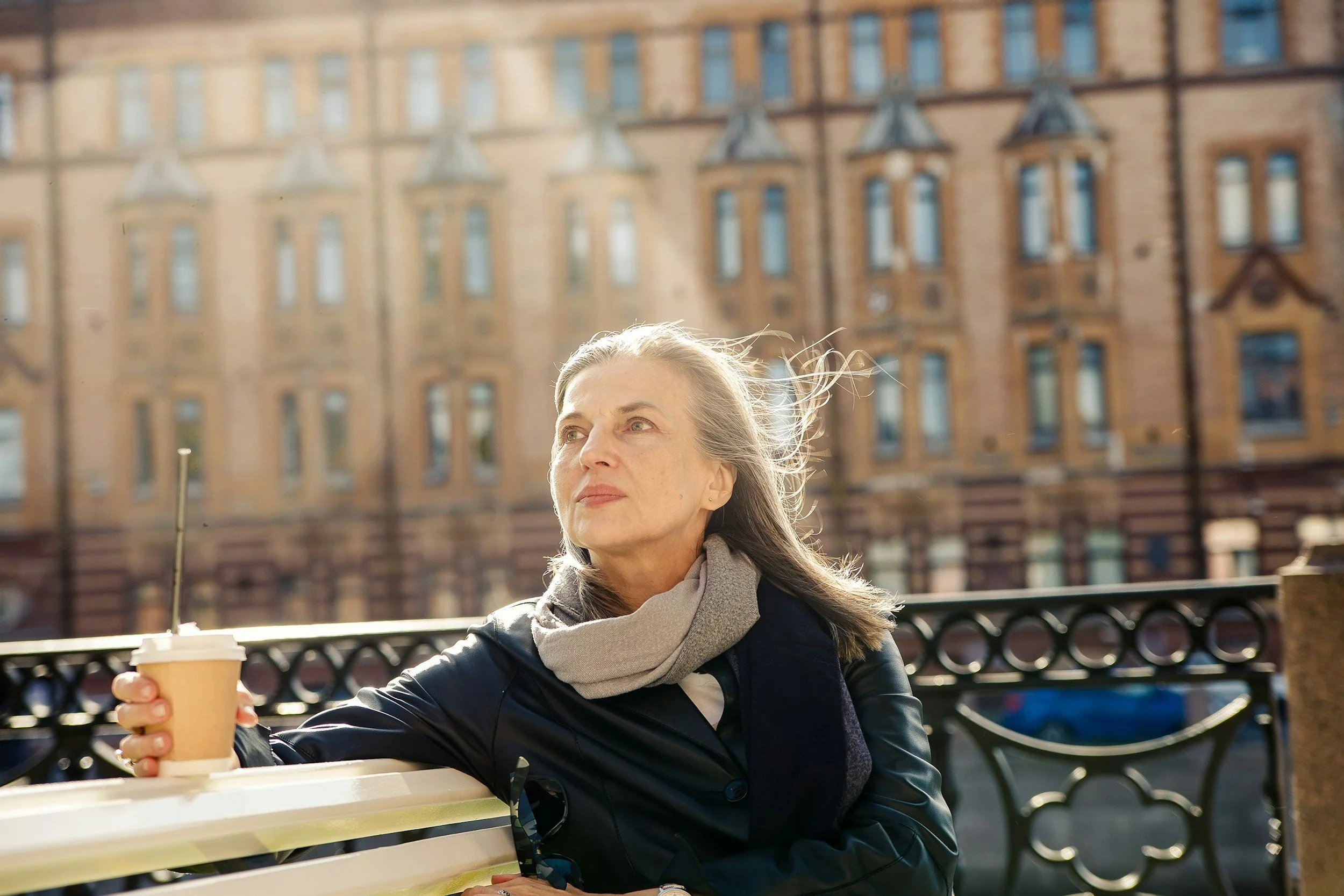 A woman sitting on a park bench holding a paper coffee cup with the cityscape in the background.