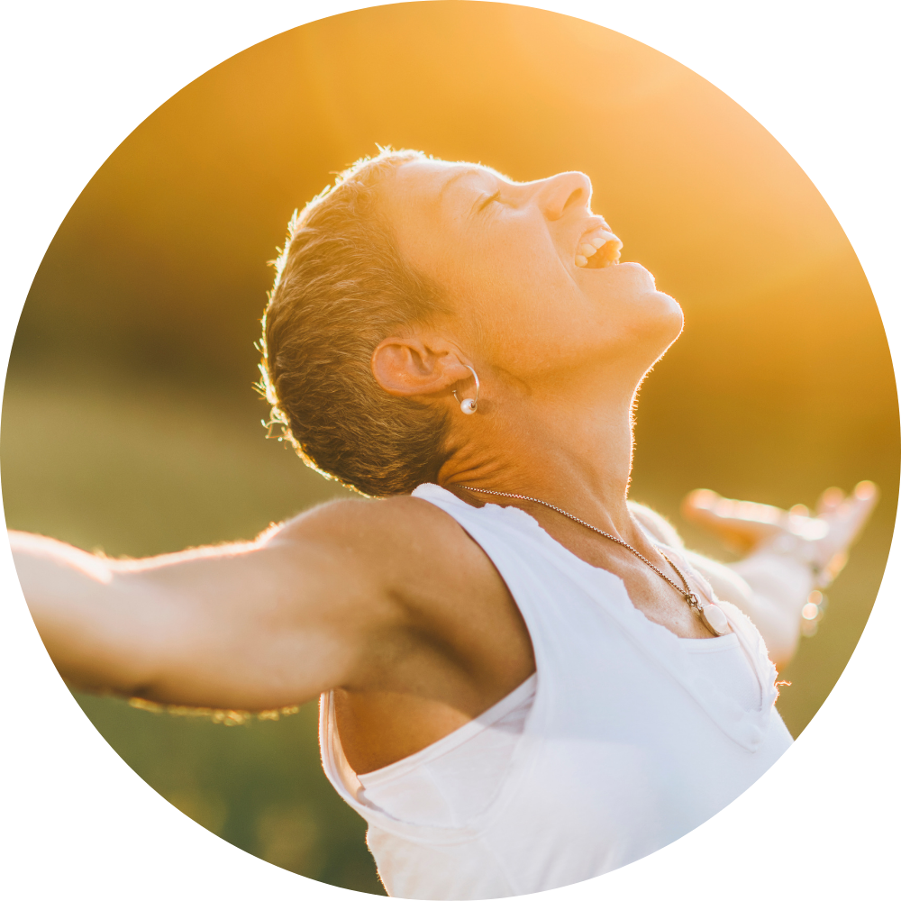 A woman with short hair, wearing a white sleeveless top and earrings, is outdoors at sunset with her arms outstretched and her face turned upward, smiling with her eyes closed.