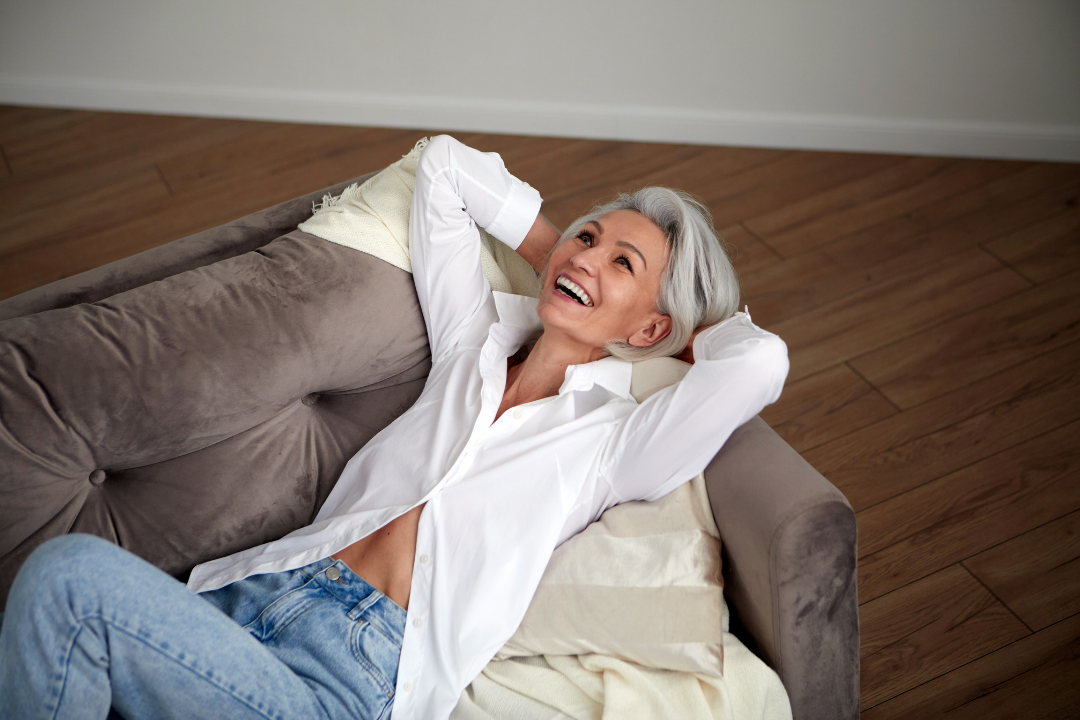 A happy older woman with gray hair lying on a brown couch, smiling and looking up, with her hands behind her head, wearing a white shirt and jeans.