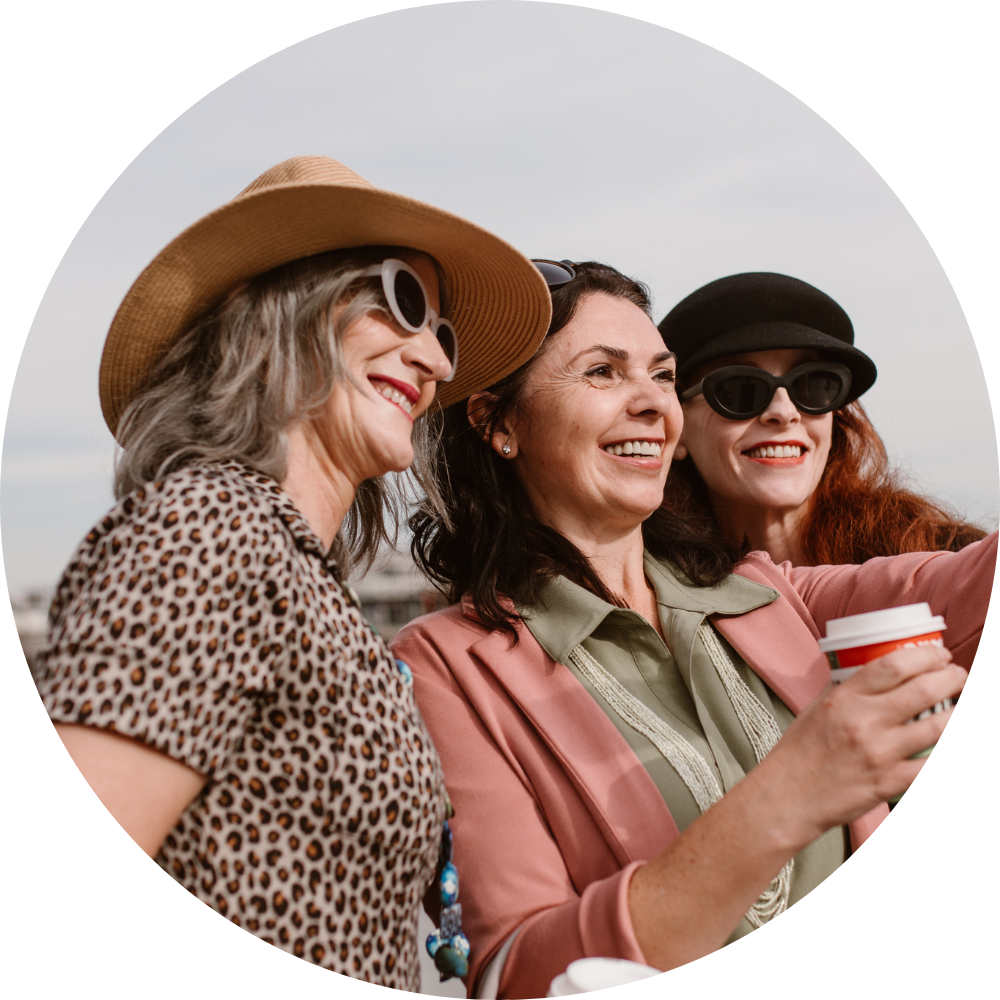 Three women smiling and taking a selfie outdoors. One woman is wearing a wide-brimmed hat and sunglasses, another is wearing sunglasses and a hat, and the third woman is holding a coffee cup.