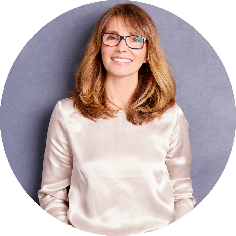 A woman with shoulder-length red hair, wearing glasses and a satin white blouse, smiling and standing against a dark purple background.