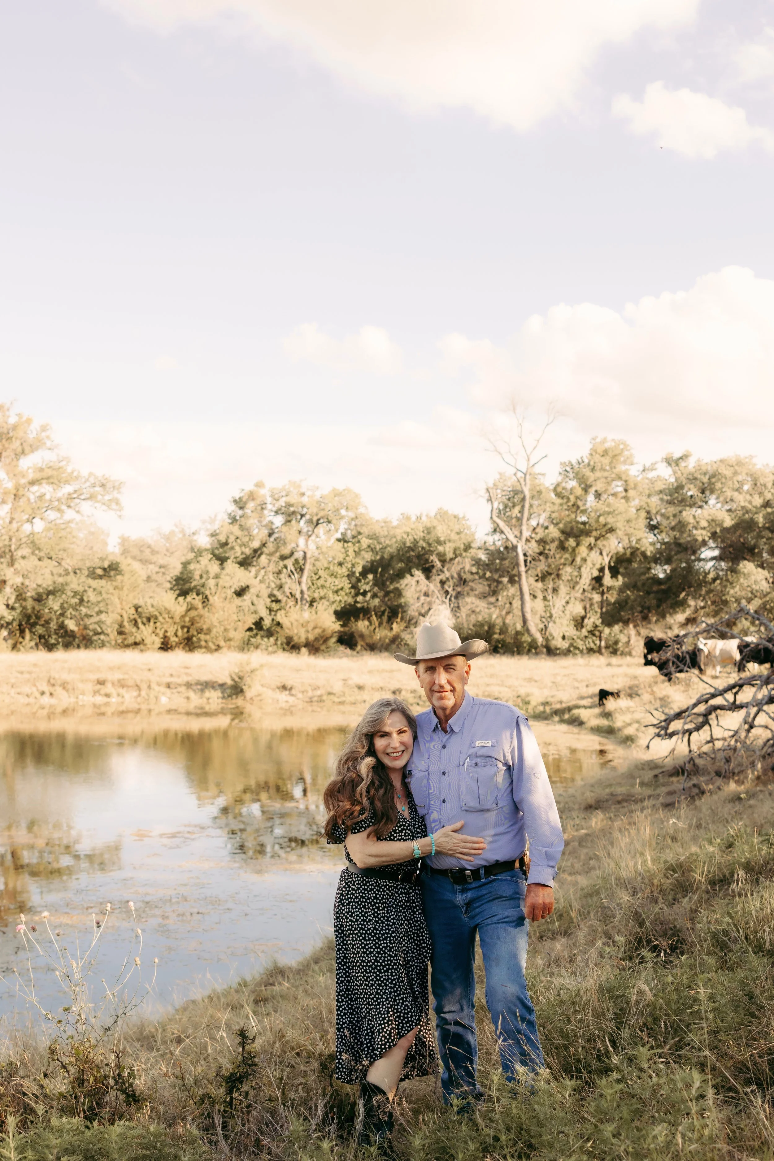 A smiling woman with long curly hair and a polka dot dress standing next to a man wearing a cowboy hat, a light blue shirt, and jeans, near a pond with trees in the background on a sunny day.
