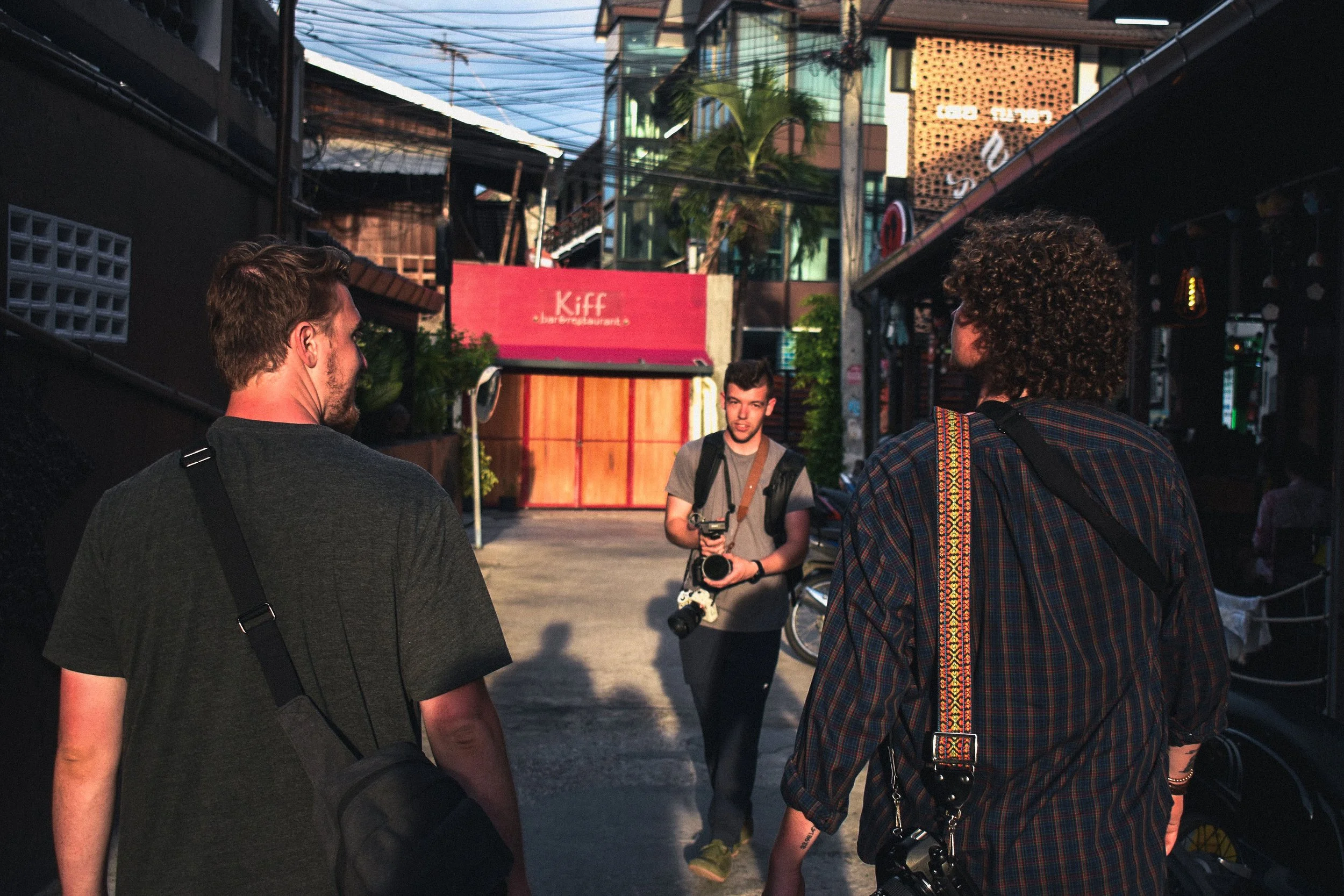 Three young men walking and talking in a narrow street or alleyway, carrying cameras and backpacks, with a red sign that reads 'Kiff' in the background.