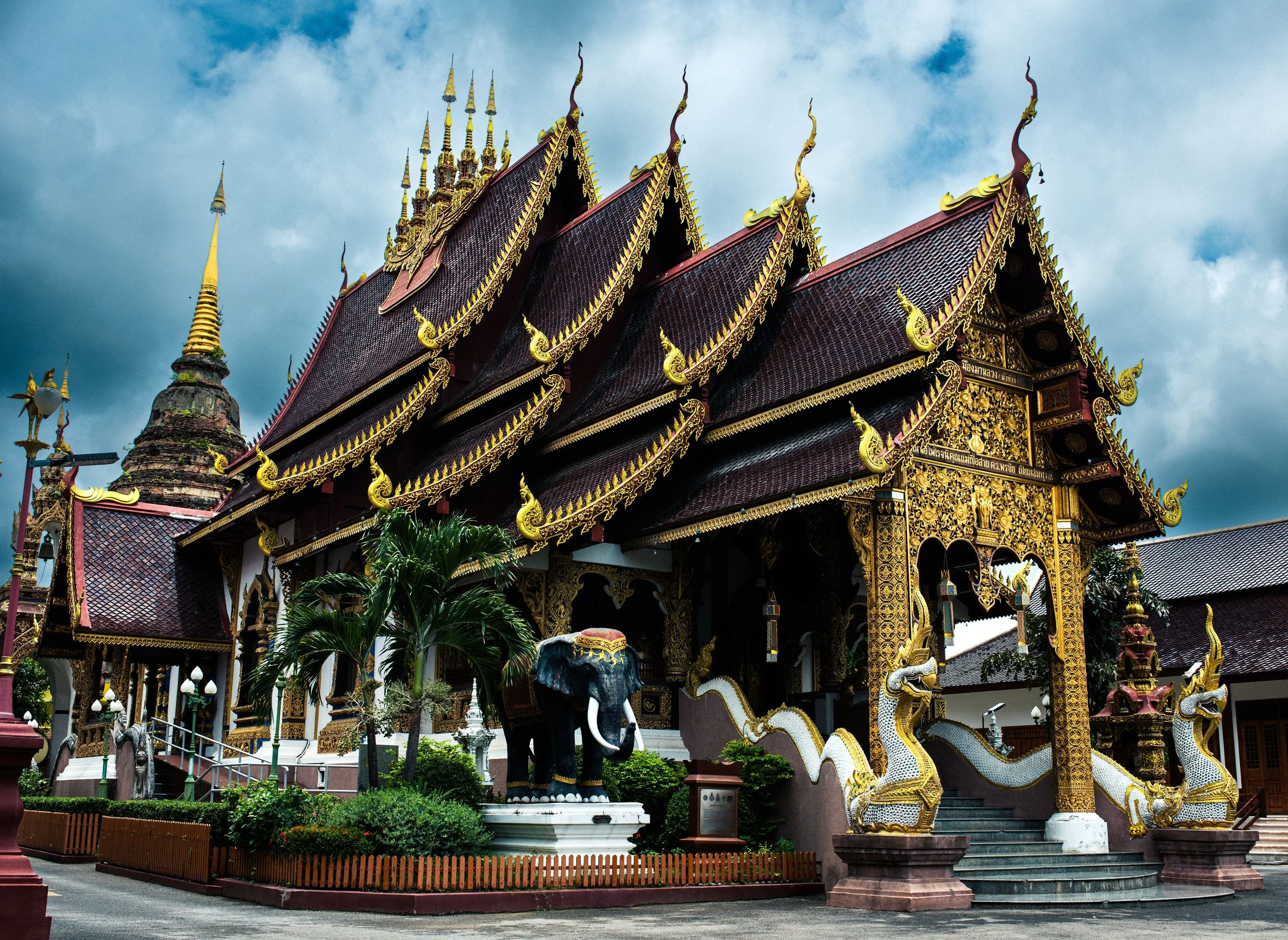 A traditional Thai temple with ornate gold detailing, dragon sculptures on stairs, and a dark cloudy sky overhead.