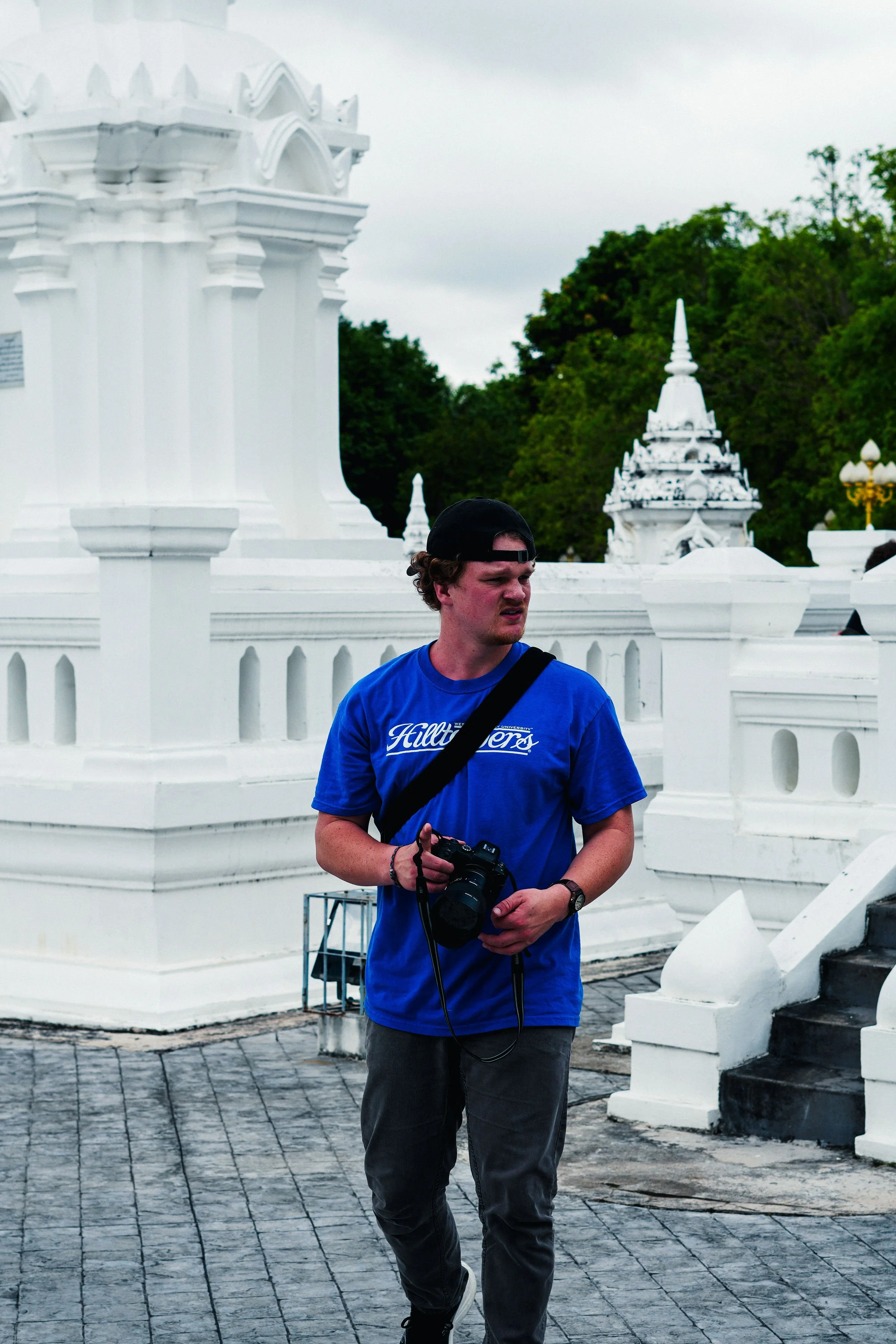 A young man in a blue t-shirt and black cap holding a camera, standing in front of white traditional Thai architecture with trees in the background.