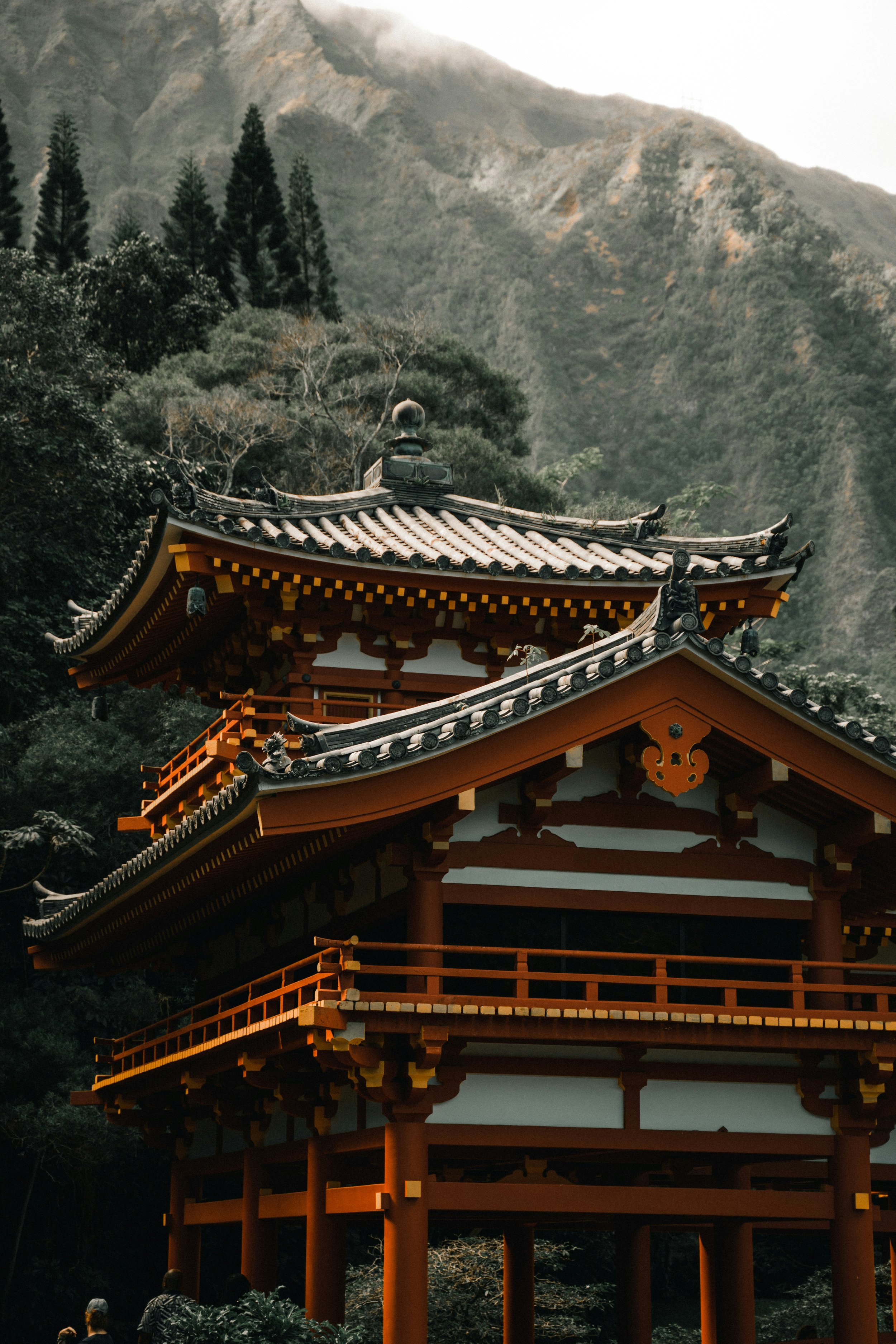 Traditional Japanese pagoda with multiple tiers, reddish-brown wooden structure, and ornate roof tiles, set against a backdrop of lush green mountains and trees.